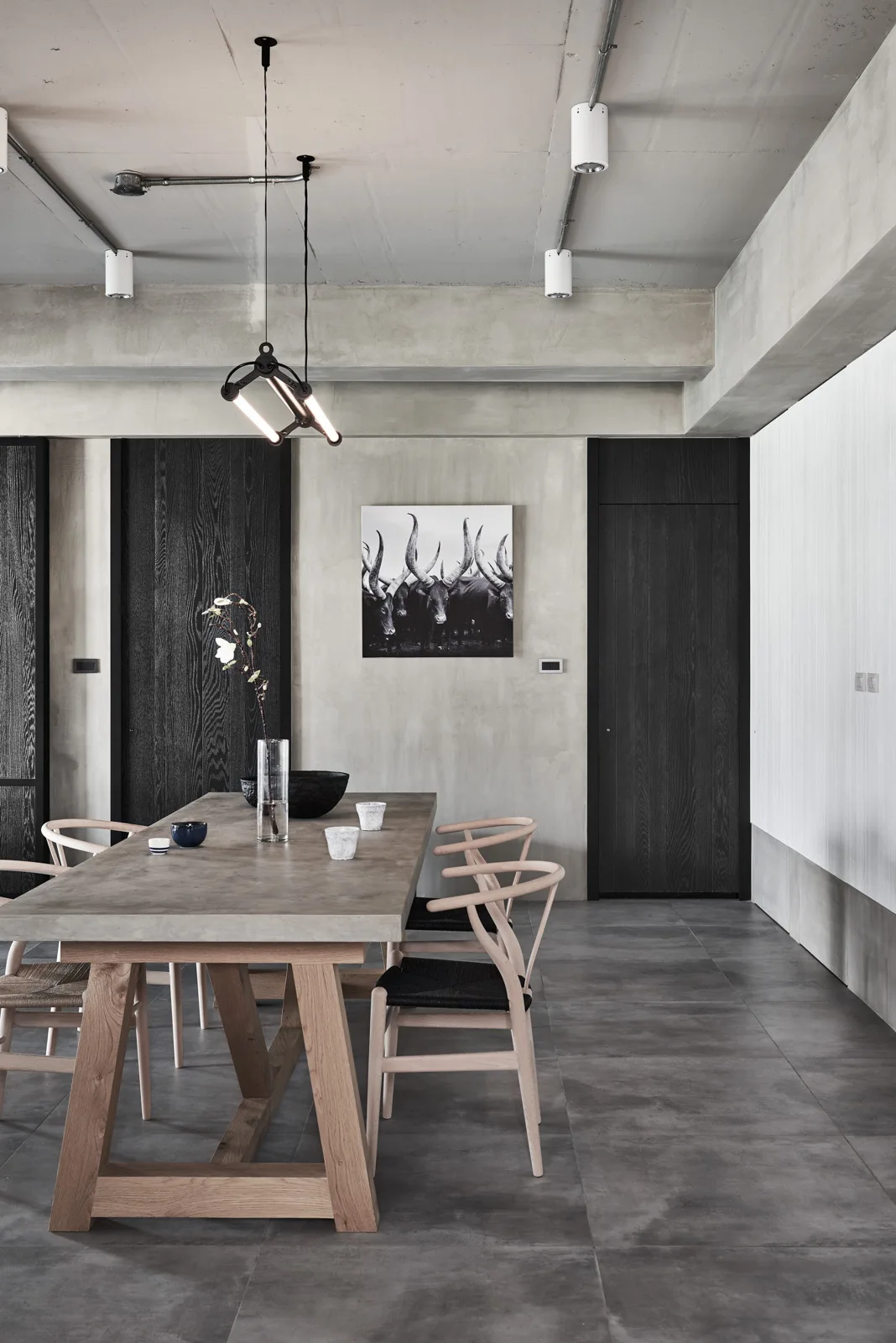 A minimalist industrial dining room features a concrete table surrounded by wooden wishbone chairs, set against a concrete ceiling and dark wooden doors.