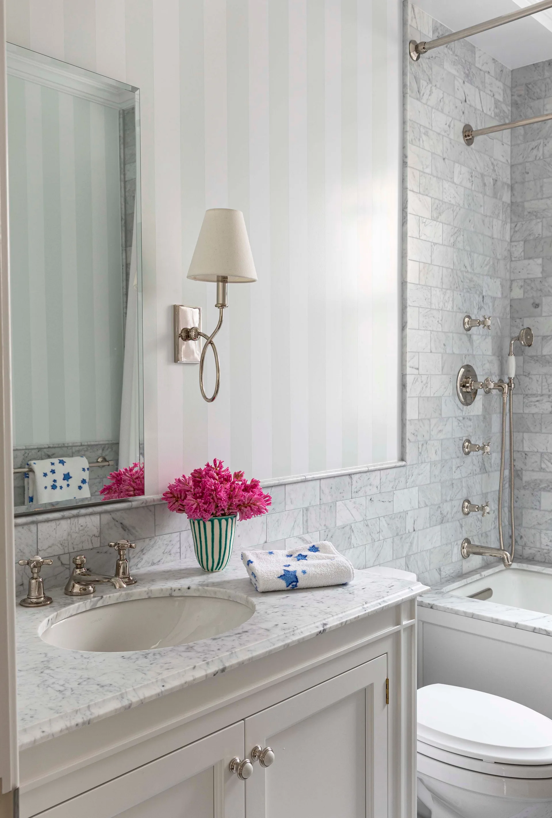 A classic bathroom vanity with a white marble countertop sits next to a tiled shower and features soft striped wallpaper.