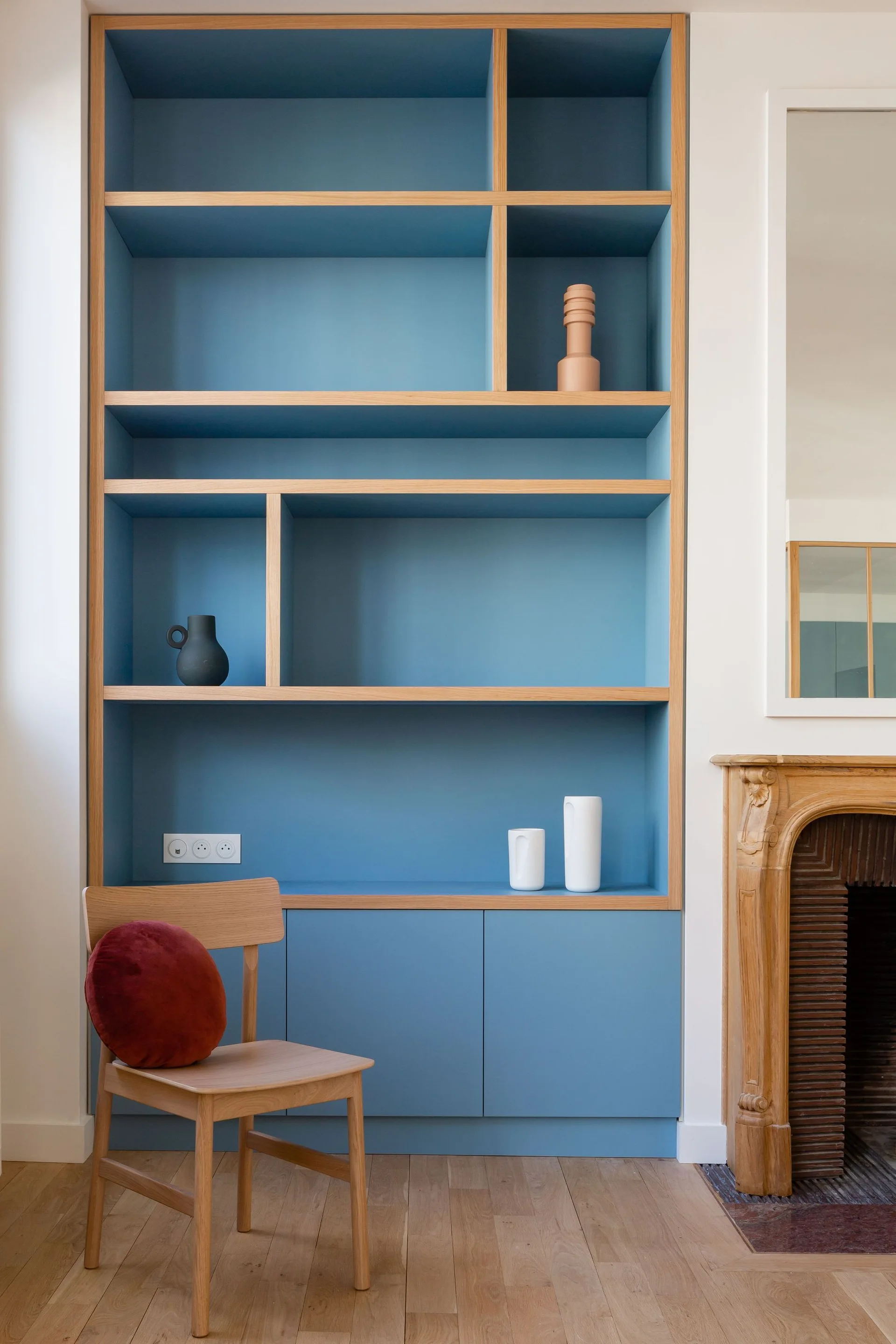 A custom blue built-in shelving unit with light wood shelves stands next to a carved wooden fireplace mantel and a simple wooden chair.