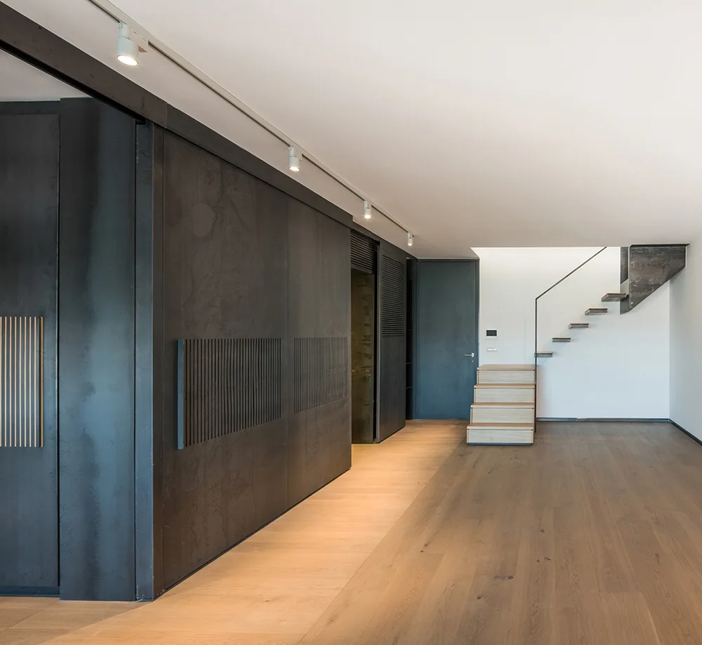 A minimalist hallway features a dark charcoal sliding storage wall, light wood flooring, and an open-riser staircase in the background.
