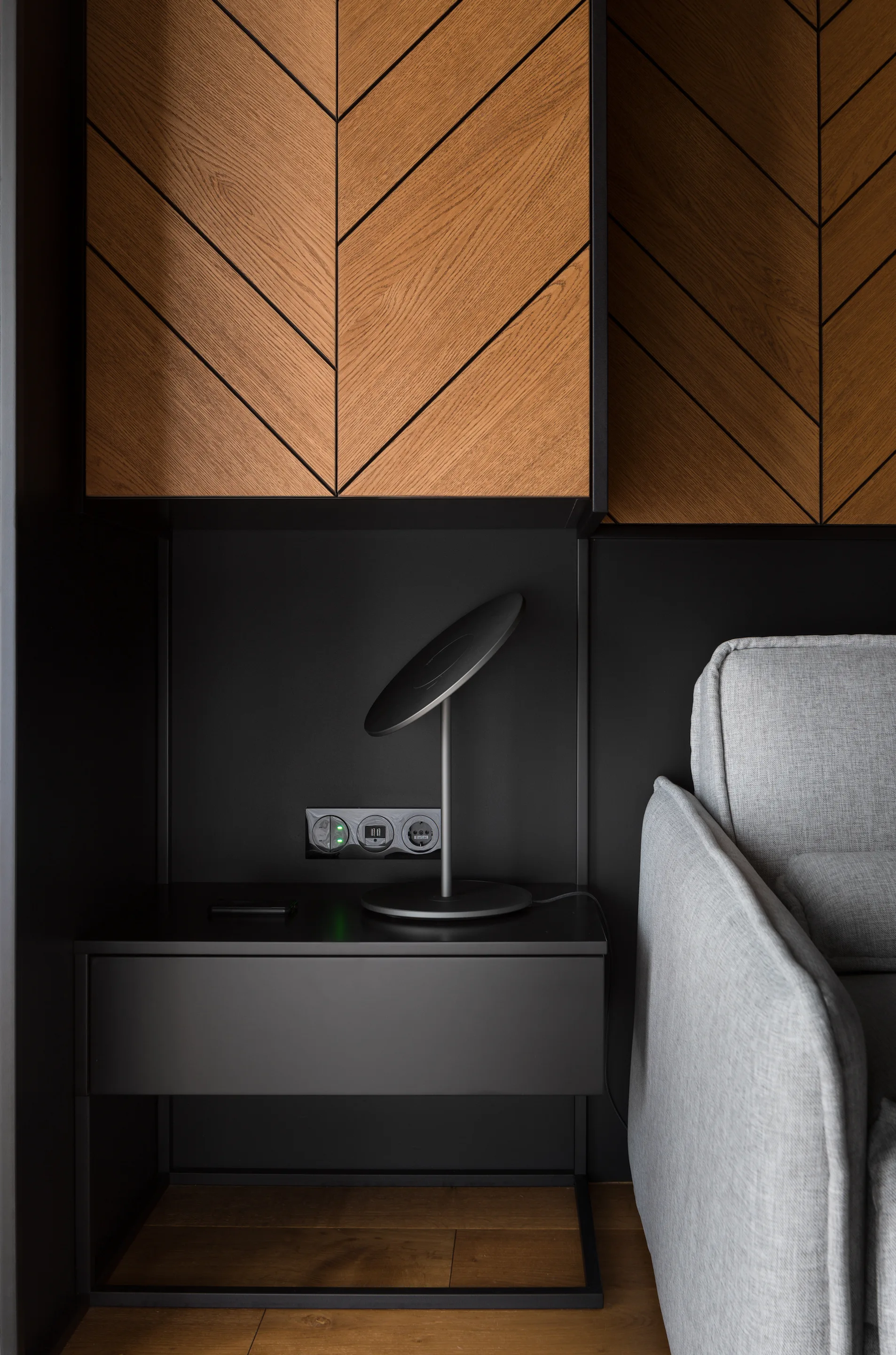 A modern bedside nook features a matte black floating nightstand beneath herringbone patterned wood wall panels next to a grey fabric sofa.