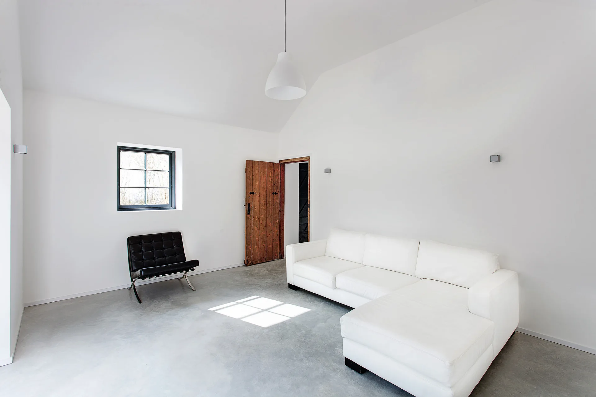 A minimalist white living room with a large white sectional sofa, a black leather armchair, and an open rustic wooden door.