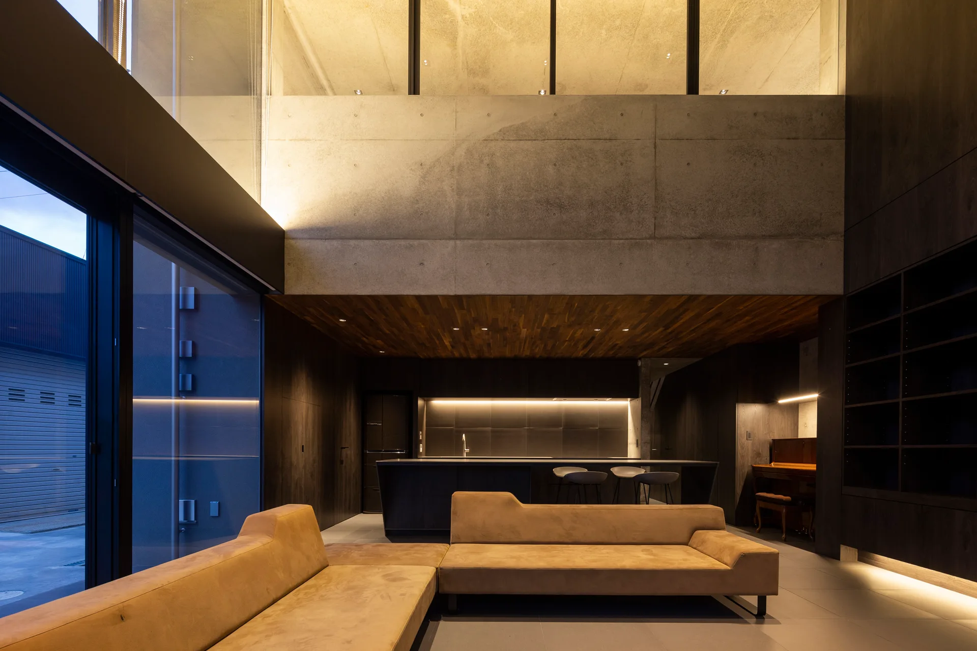 A modern open-concept living area with a large tan sectional sofa, exposed concrete walls, and dark wood cabinetry in the kitchen background.
