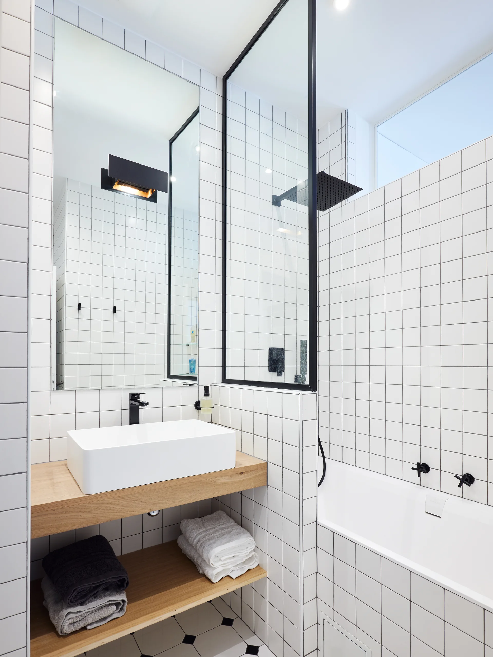 A contemporary bathroom with white square grid tiles, a floating wood vanity shelf, a white vessel sink, and a black-framed glass partition.