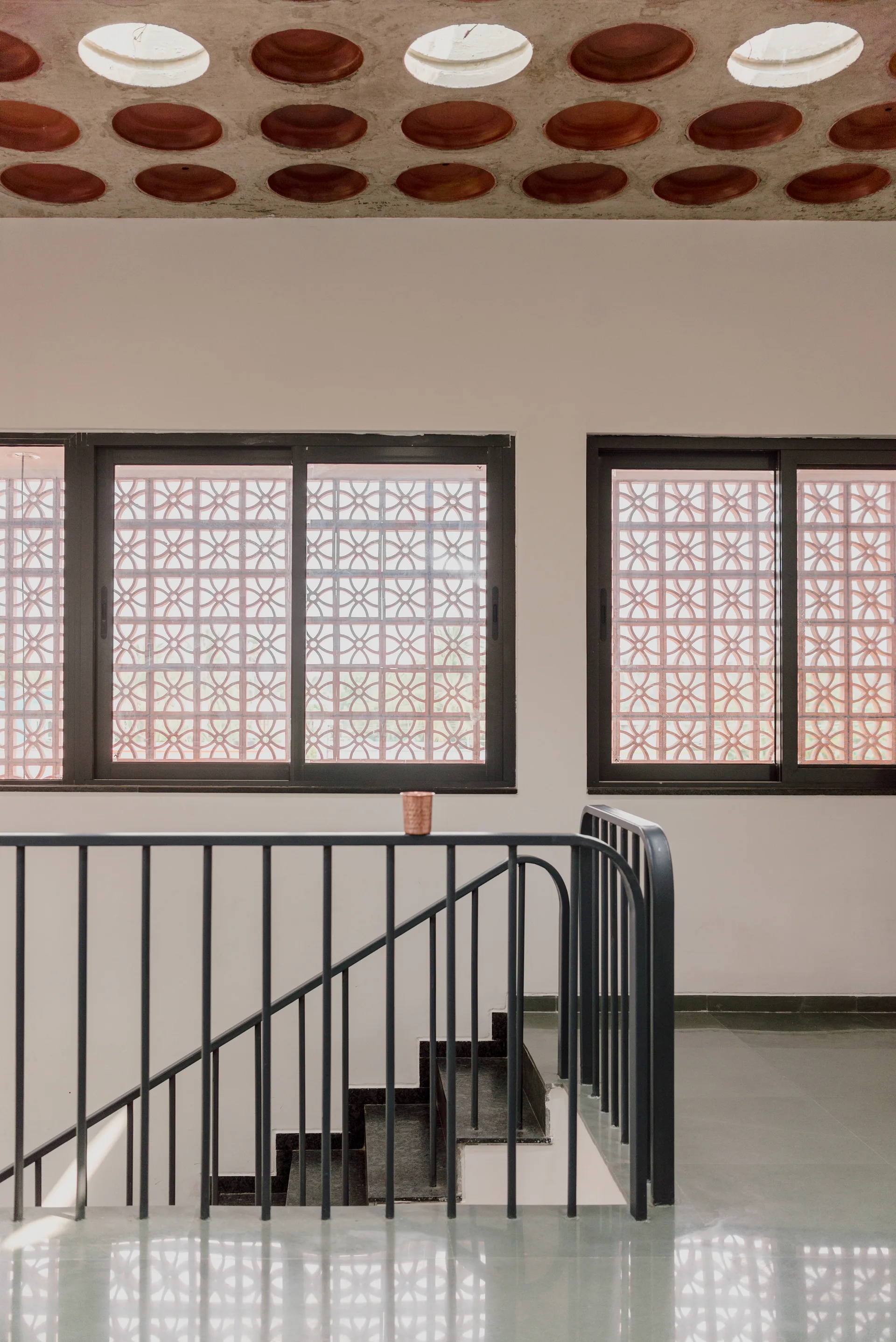A black metal staircase railing stands on a grey stone floor in front of white walls with two windows featuring patterned terracotta screens.