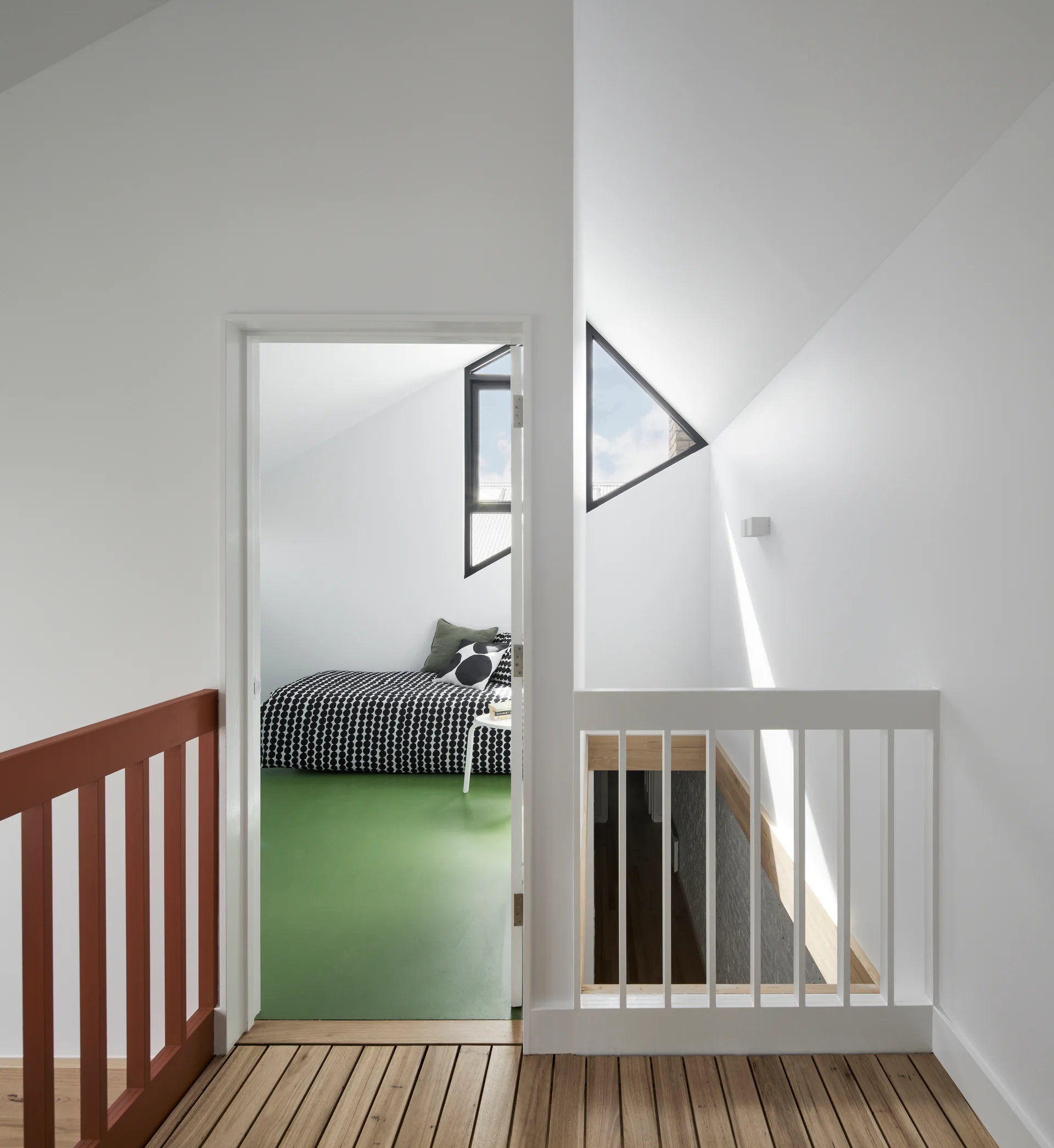 A view from a slatted wood landing with a white stairwell railing looking into a bedroom with a green floor, patterned bed, and triangular window.