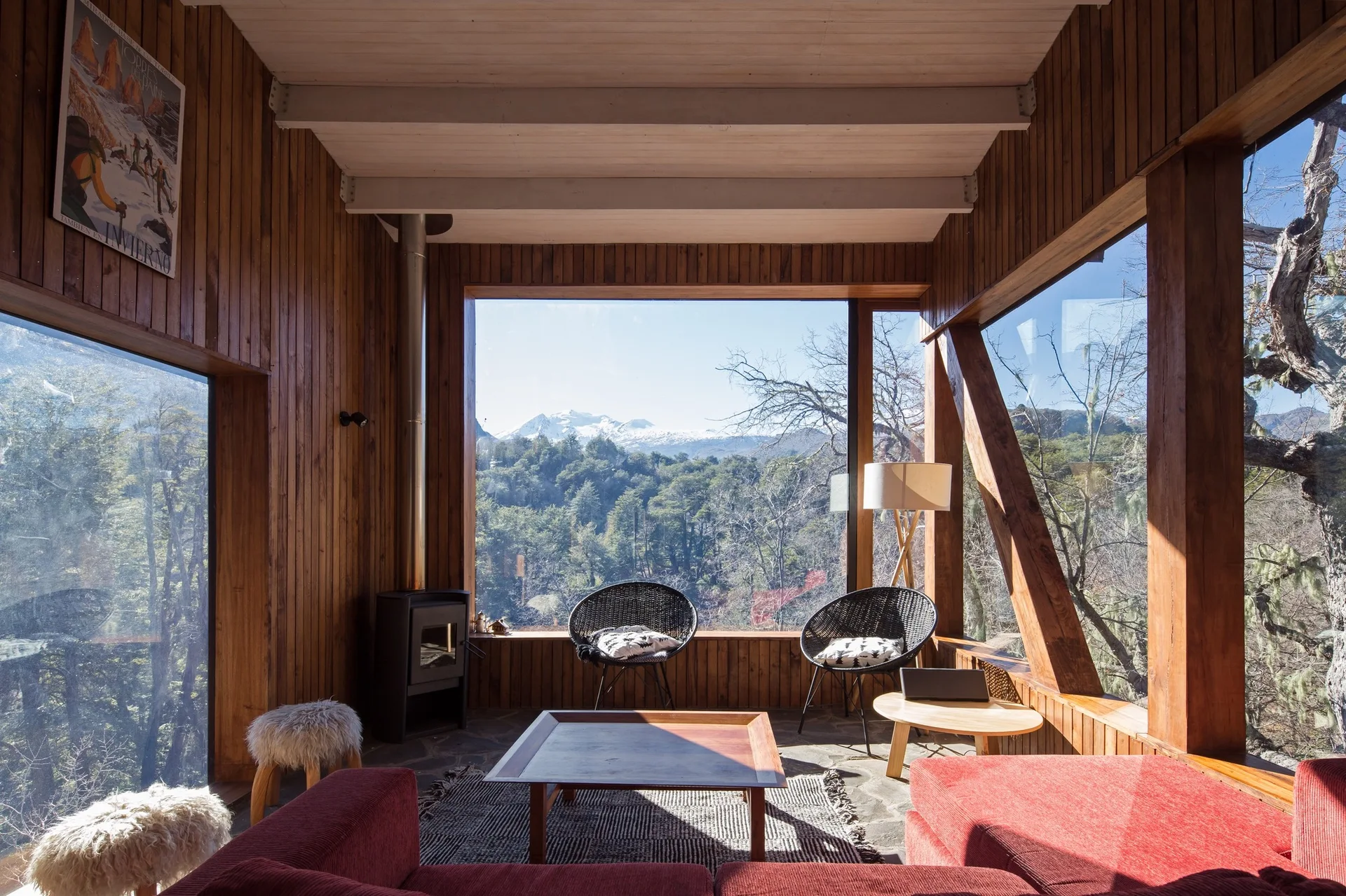 A rustic cabin living room features vertical wood paneling, a large red sofa, stone tile flooring, and a large picture window framing mountain views.