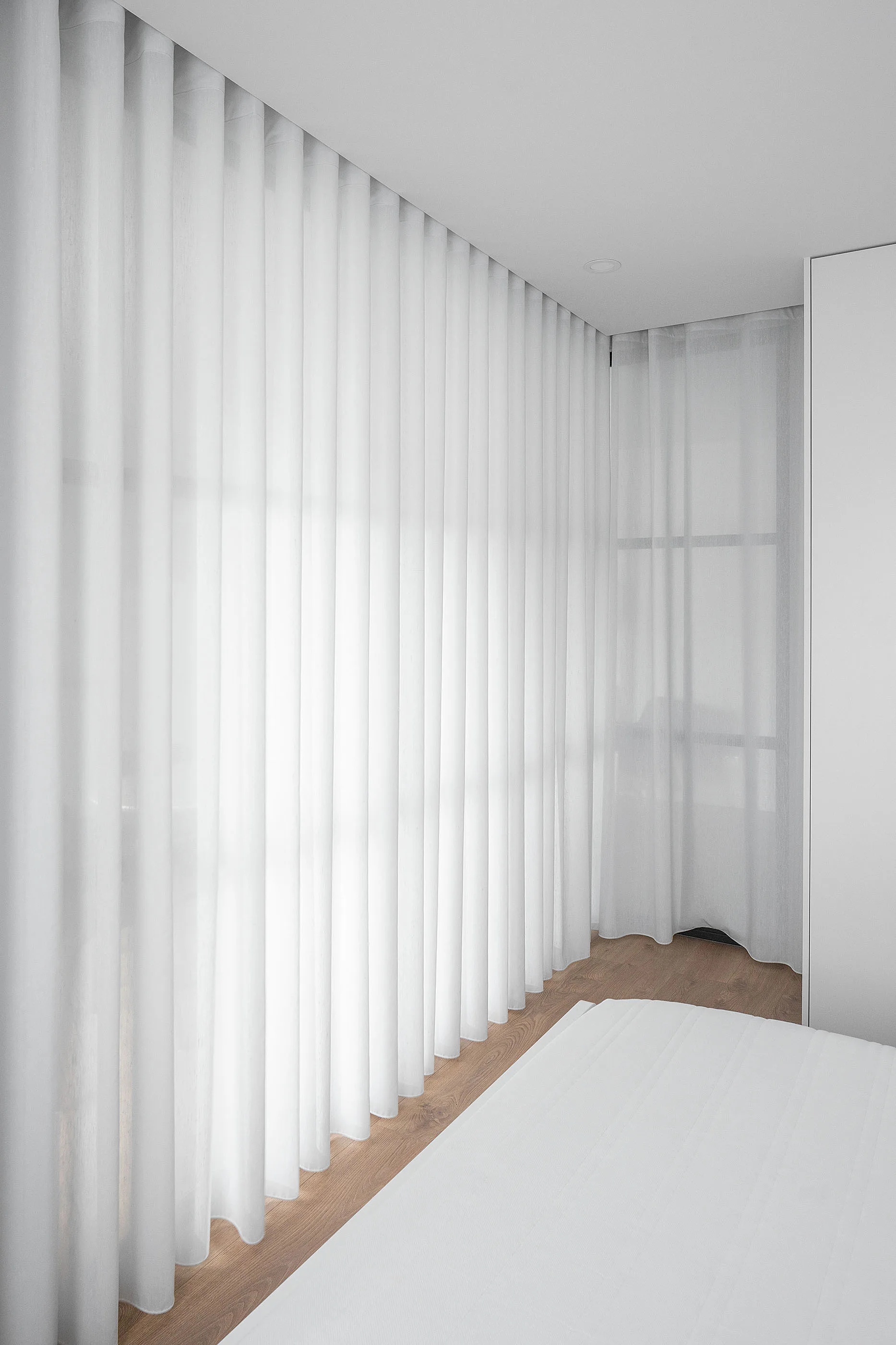 White floor-to-ceiling sheer curtains cover a corner window in a minimalist bedroom with light wood flooring.