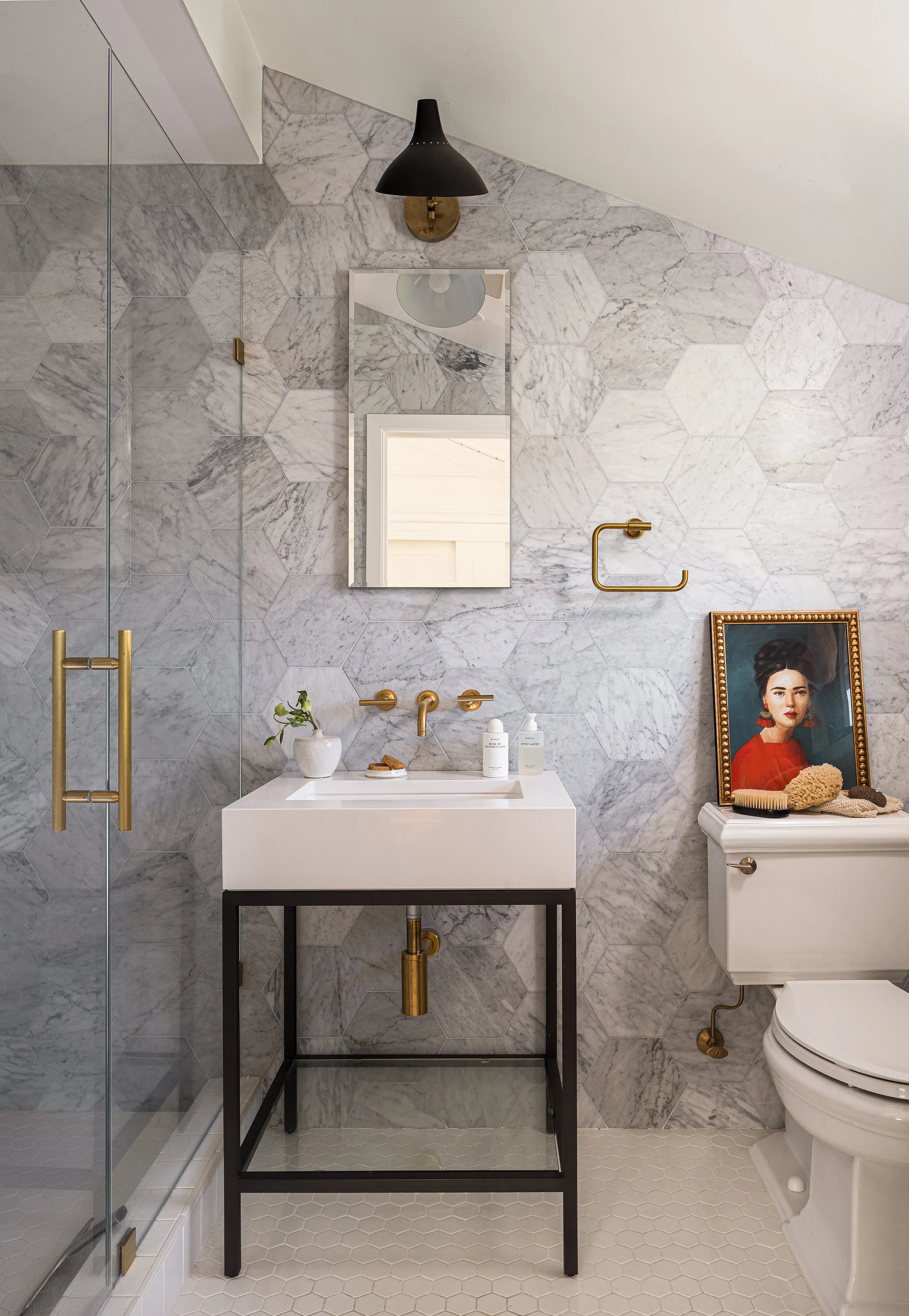 A bathroom featuring a white console sink with a black frame, hexagonal marble wall tiles, brushed brass hardware, and a glass shower door.