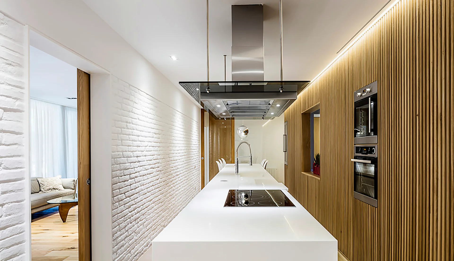 A modern kitchen featuring a long white island with a built-in cooktop, a slatted wood wall with integrated ovens, and a painted white brick wall.