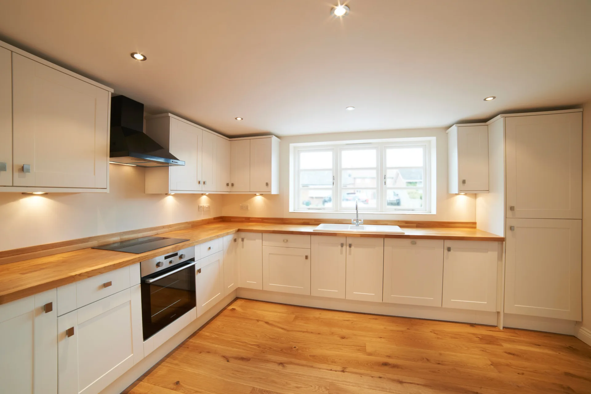 An L-shaped kitchen featuring white shaker cabinets, warm wood butcher block countertops, a centered window, and wood flooring.