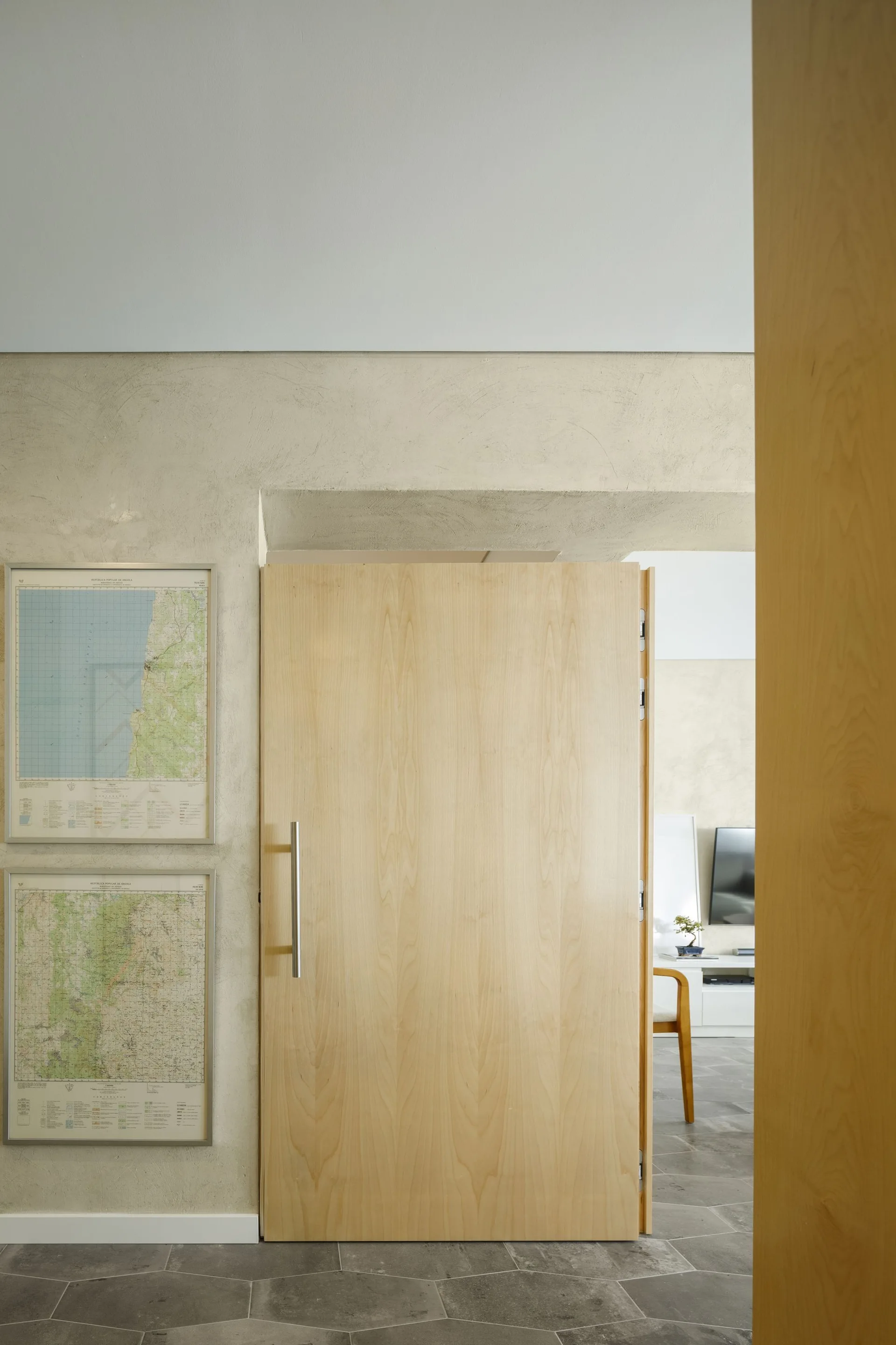 A minimalist hallway features textured beige plaster walls, framed maps, grey hexagonal floor tiles, and an open light wood door leading to a living room.