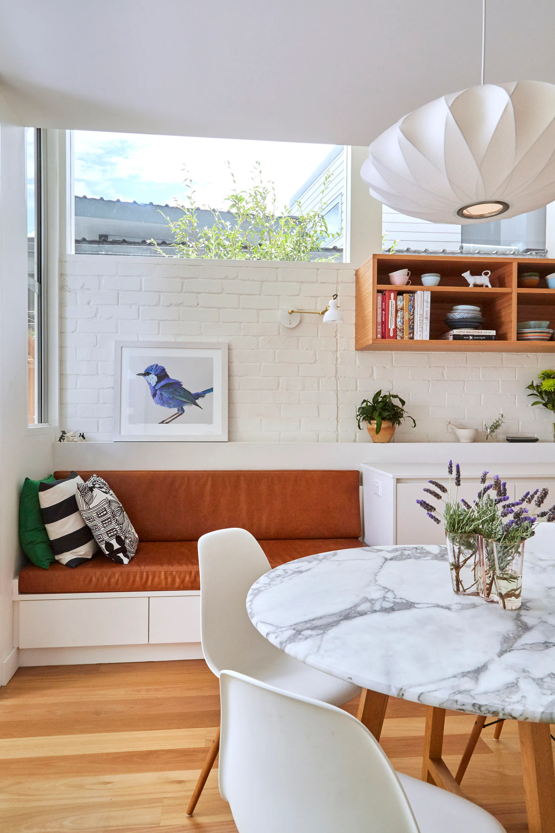 A bright dining nook features a marble table, white molded chairs, and a built-in banquette with a brown leather cushion against a white brick wall.