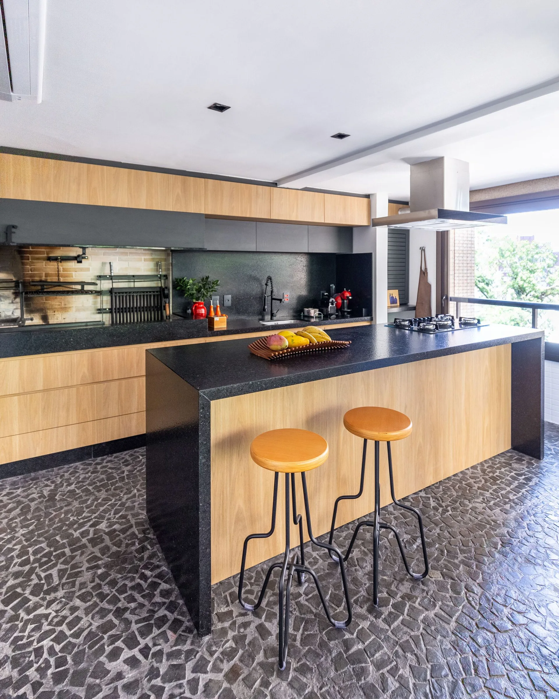 A modern kitchen featuring natural wood cabinets, dark stone countertops, a central island with stools, and a grey mosaic stone floor.