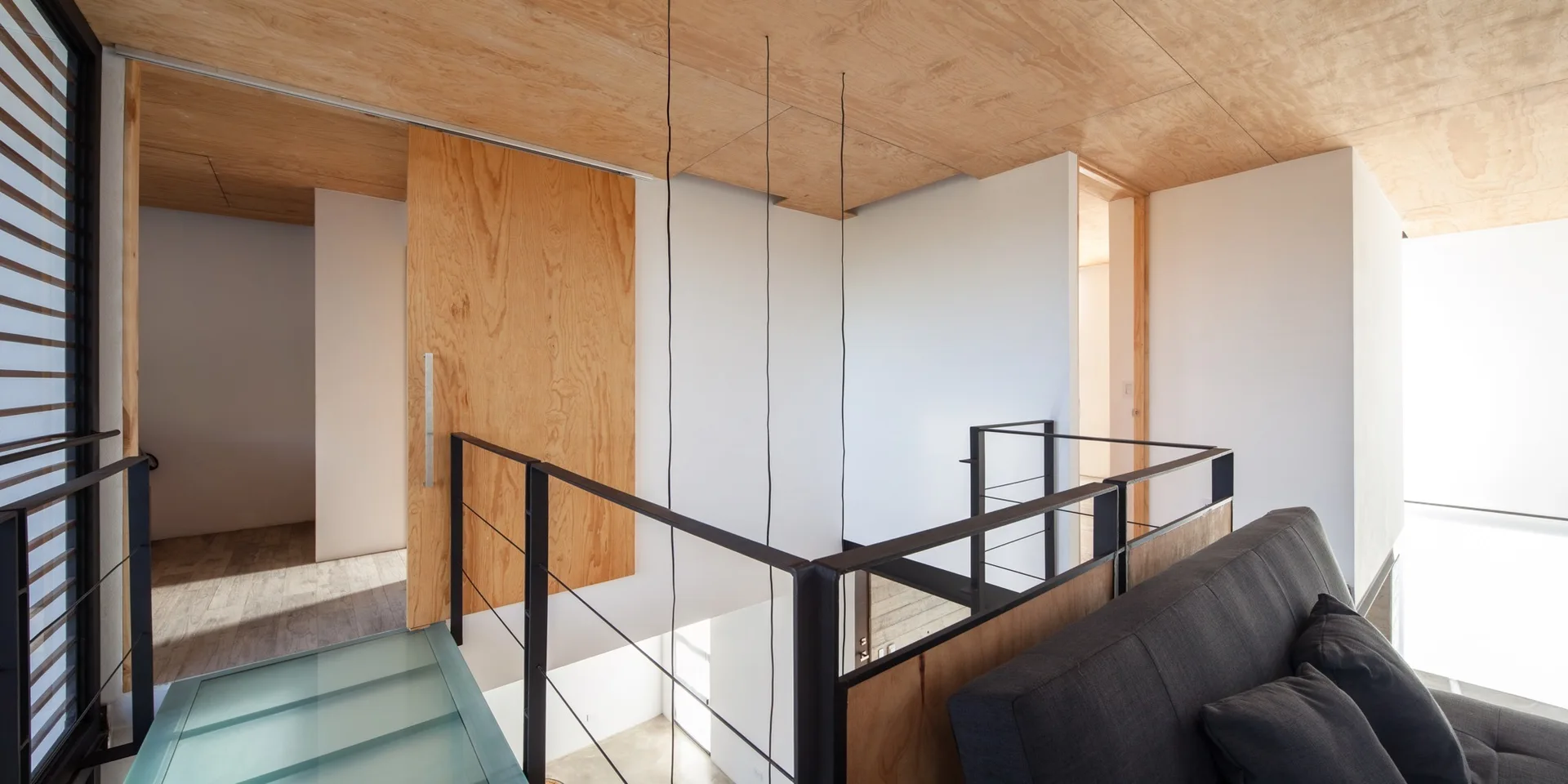 A minimalist loft landing features a plywood paneled ceiling, a black metal safety railing, a dark grey sofa, and a natural wood sliding door.