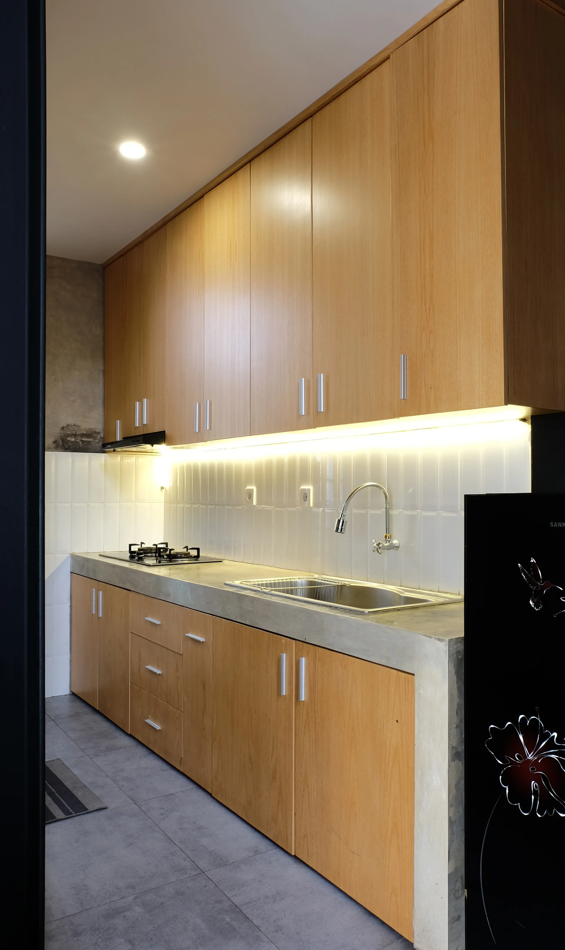 A modern linear kitchen with light wood cabinets, a concrete countertop with a sink, and vertically stacked white subway tiles illuminated by LED lights.