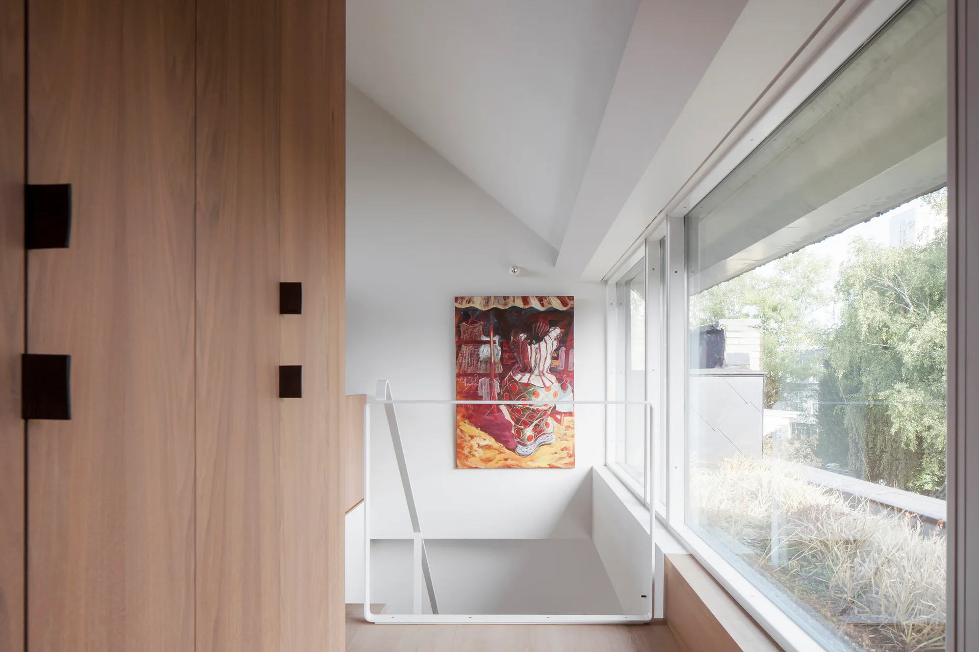 A minimalist staircase landing features warm wood paneling, a white metal railing, and a vibrant painting under a sloped ceiling next to a large window.