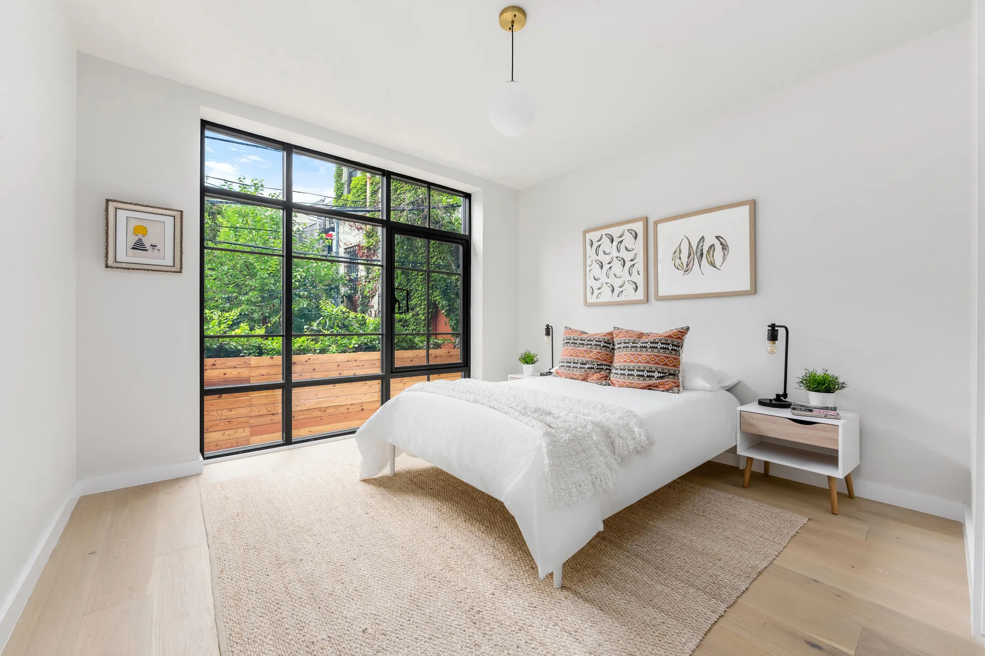 A modern white bedroom features a black-framed grid window showing leafy trees, a white bed, and a natural fiber area rug.