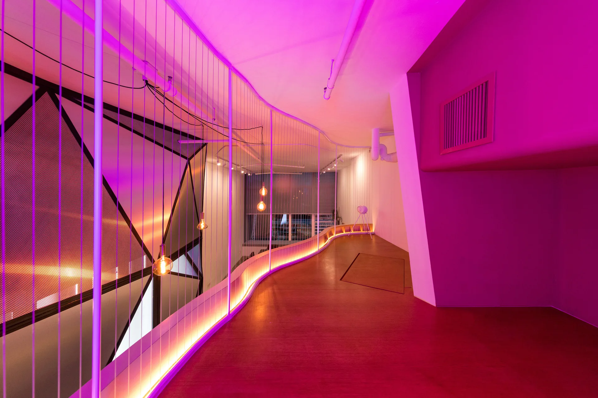 A modern curved hallway illuminated by pink light features a vertical cable railing system, warm pendant bulb lights, and wood-toned flooring.