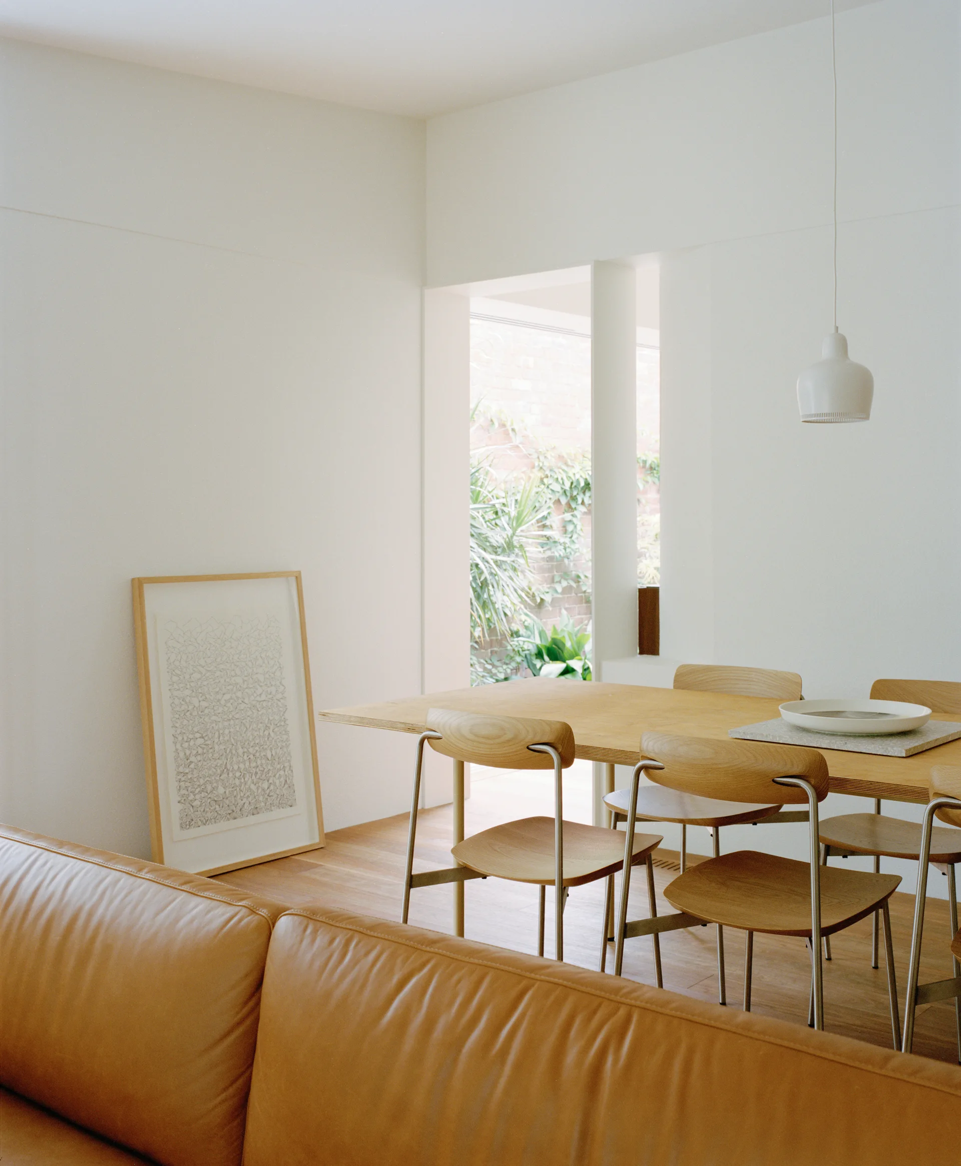 A light wood dining table and chairs are set on a wood floor next to a white wall with leaning framed artwork and an opening to a green courtyard.