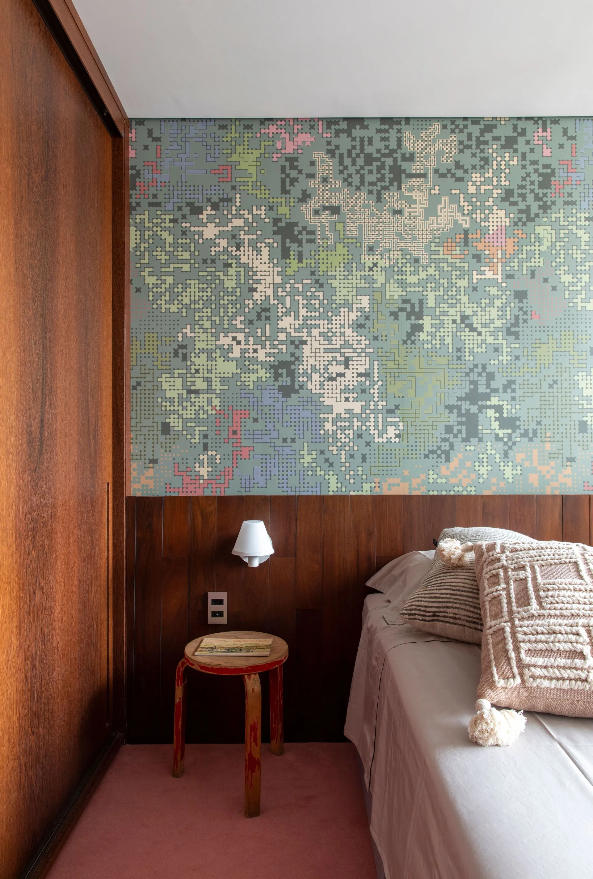 A bedroom corner features a bed with textured cushions next to wood wall paneling, multi-colored pixelated wallpaper, and a wooden stool.