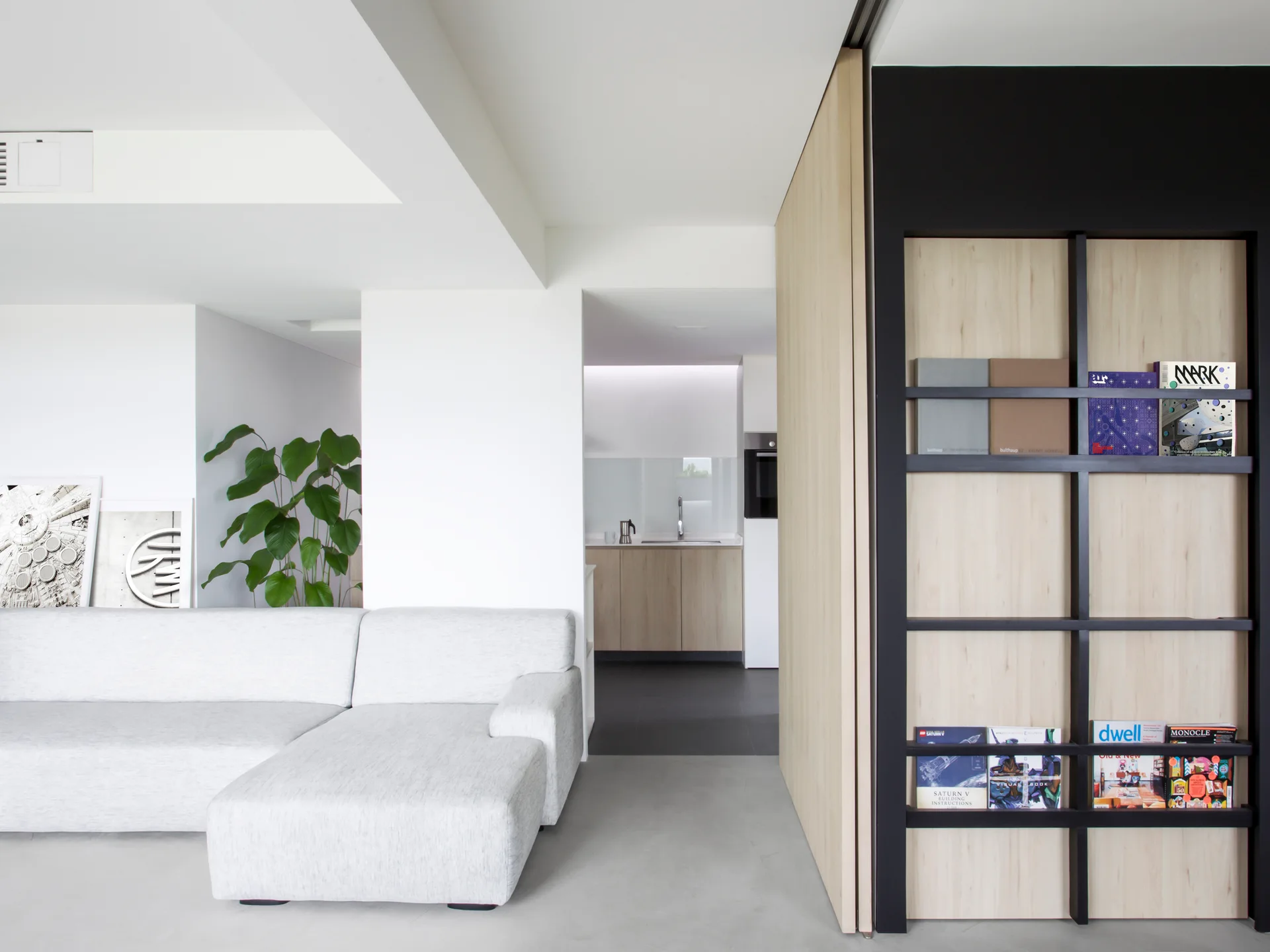 A minimalist living room features a light grey sectional sofa and a built-in light wood shelf with a black frame displaying books.