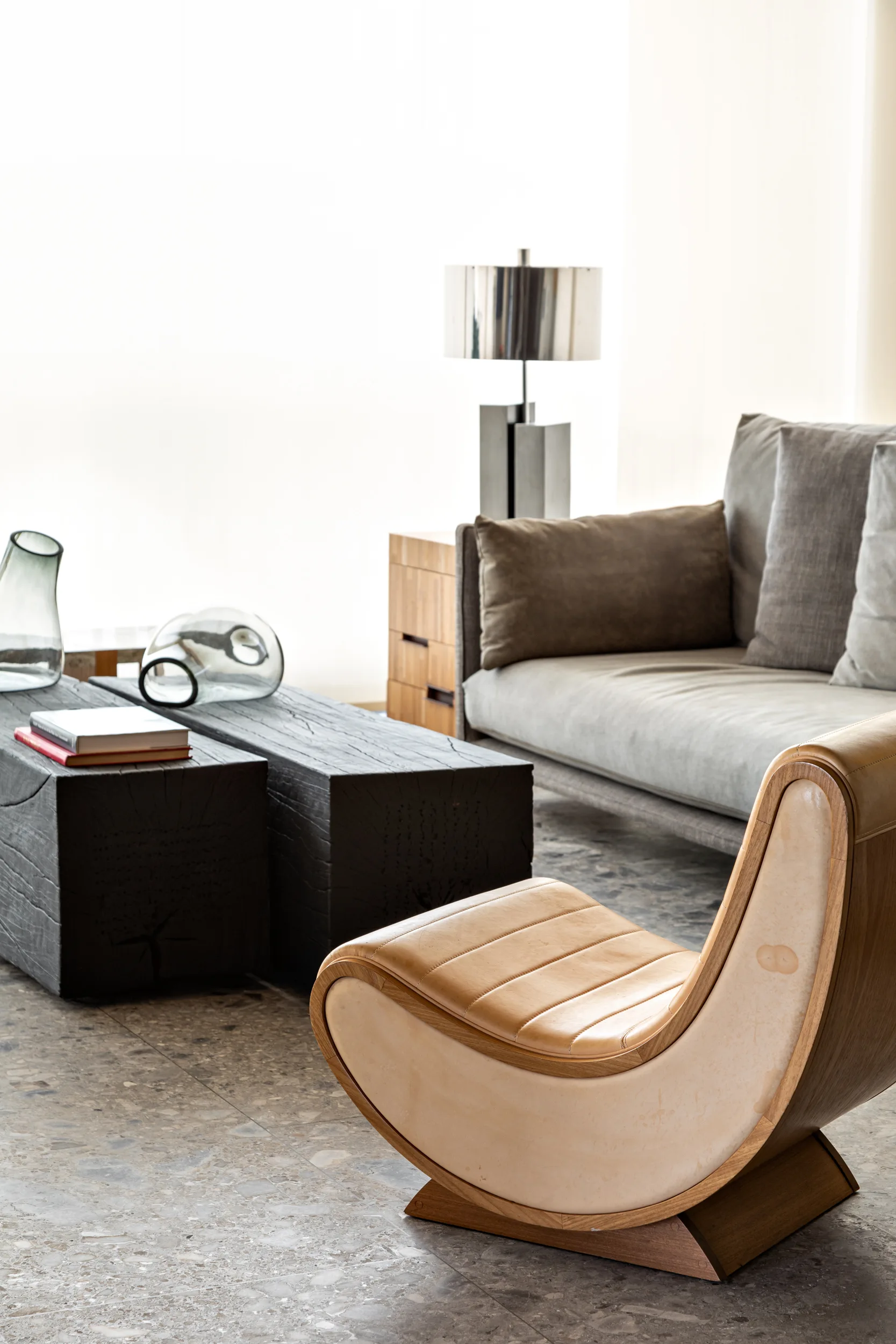 A contemporary living room features a curved tan leather accent chair next to a low charred wood coffee table and a grey fabric sofa.