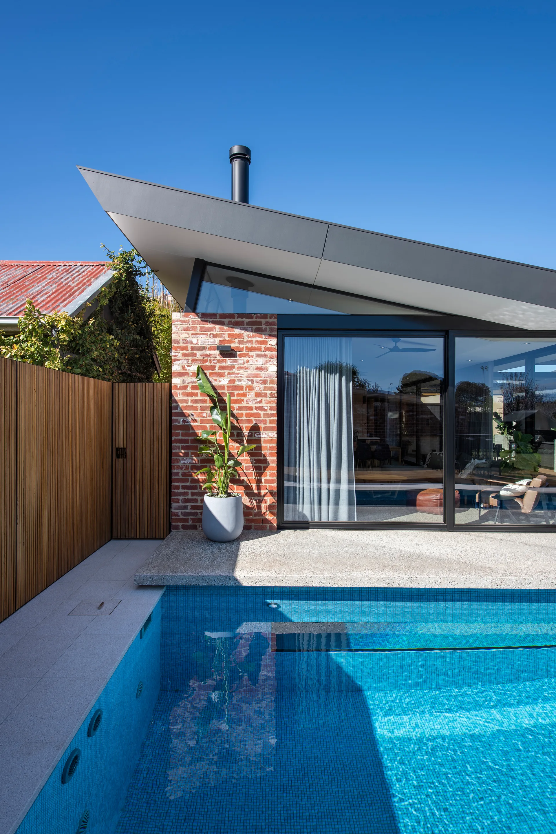A modern backyard swimming pool next to a red brick house wall, featuring a sloped dark grey roof and a vertical wood slat fence under a clear blue sky.