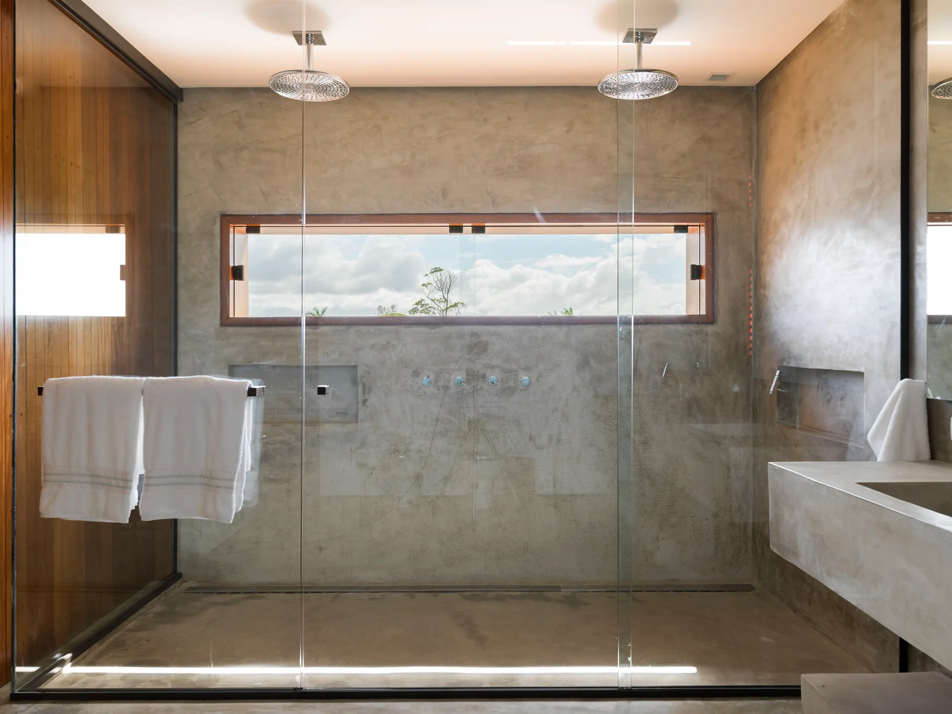 A modern bathroom featuring a spacious walk-in shower with concrete walls, two ceiling-mounted shower heads, and a long horizontal window.