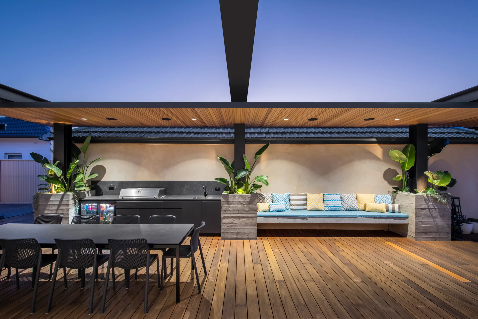 A modern outdoor deck features a wood-paneled ceiling, a black kitchen suite, a long concrete bench with colorful pillows, and a black dining table.
