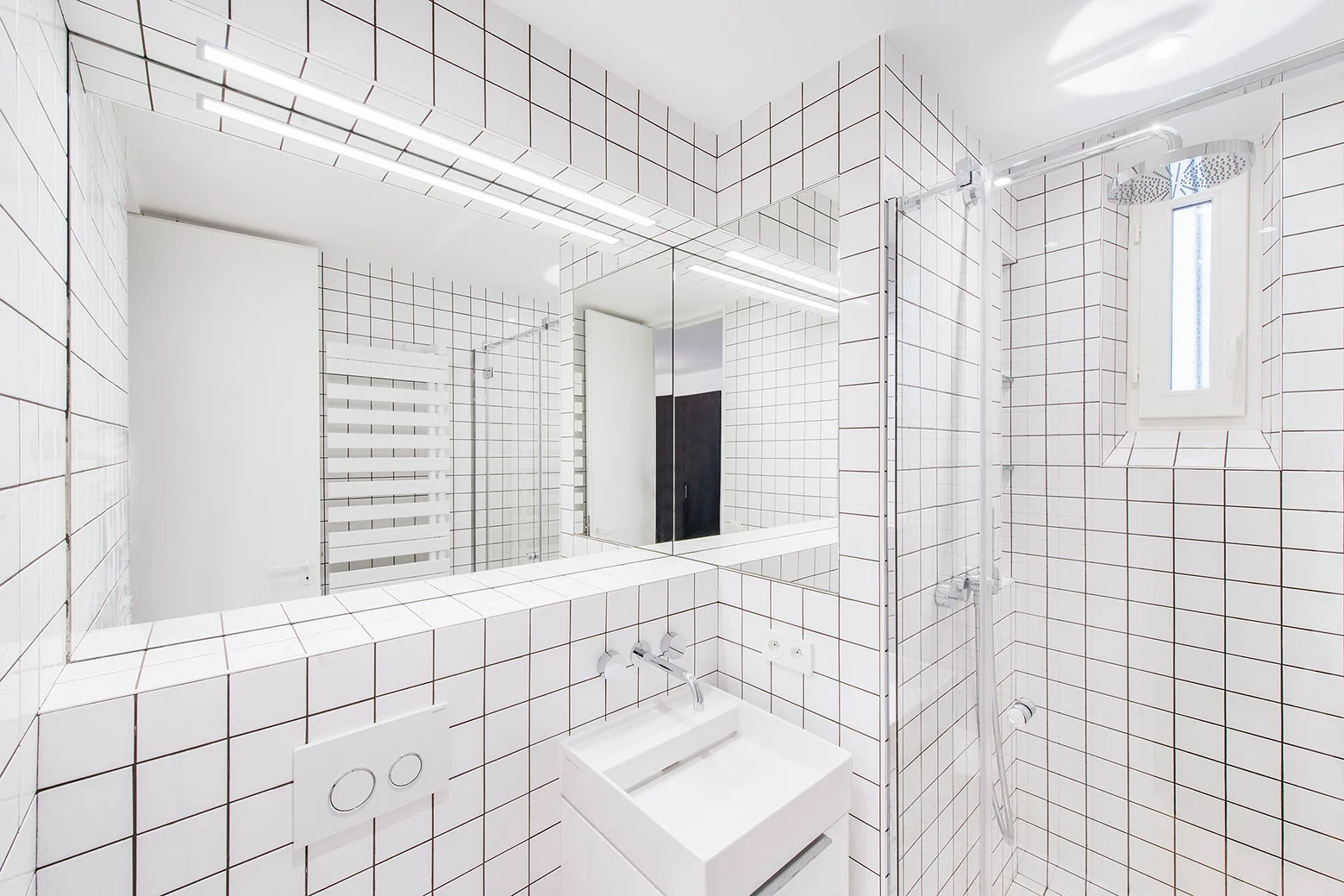 A minimalist bathroom with white square tiles and black grout covering the walls, featuring a wall-mounted sink and a large rectangular mirror.