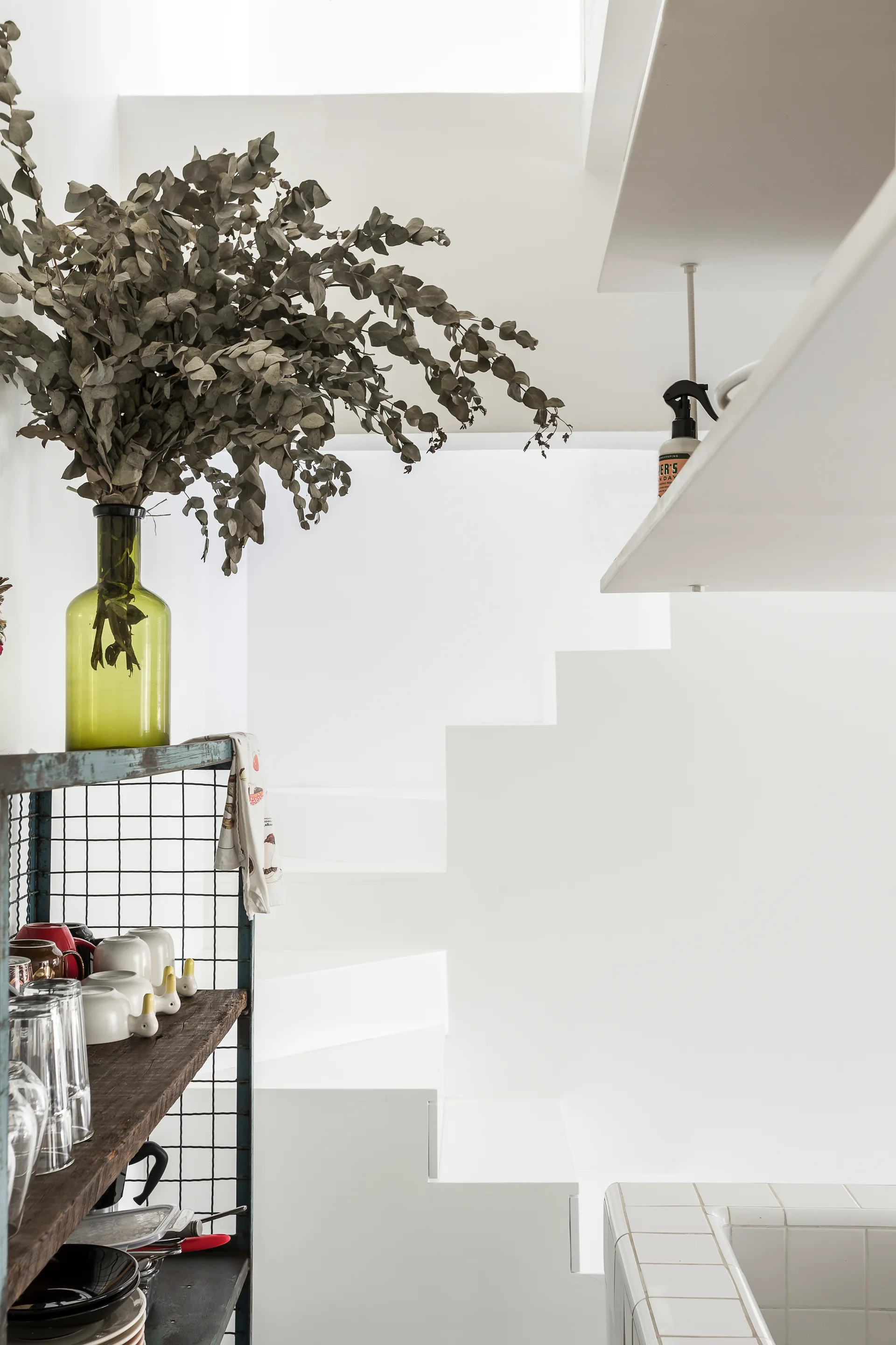 A minimalist white staircase is visible behind a metal wire shelving unit holding glassware and a green vase with dried eucalyptus branches.