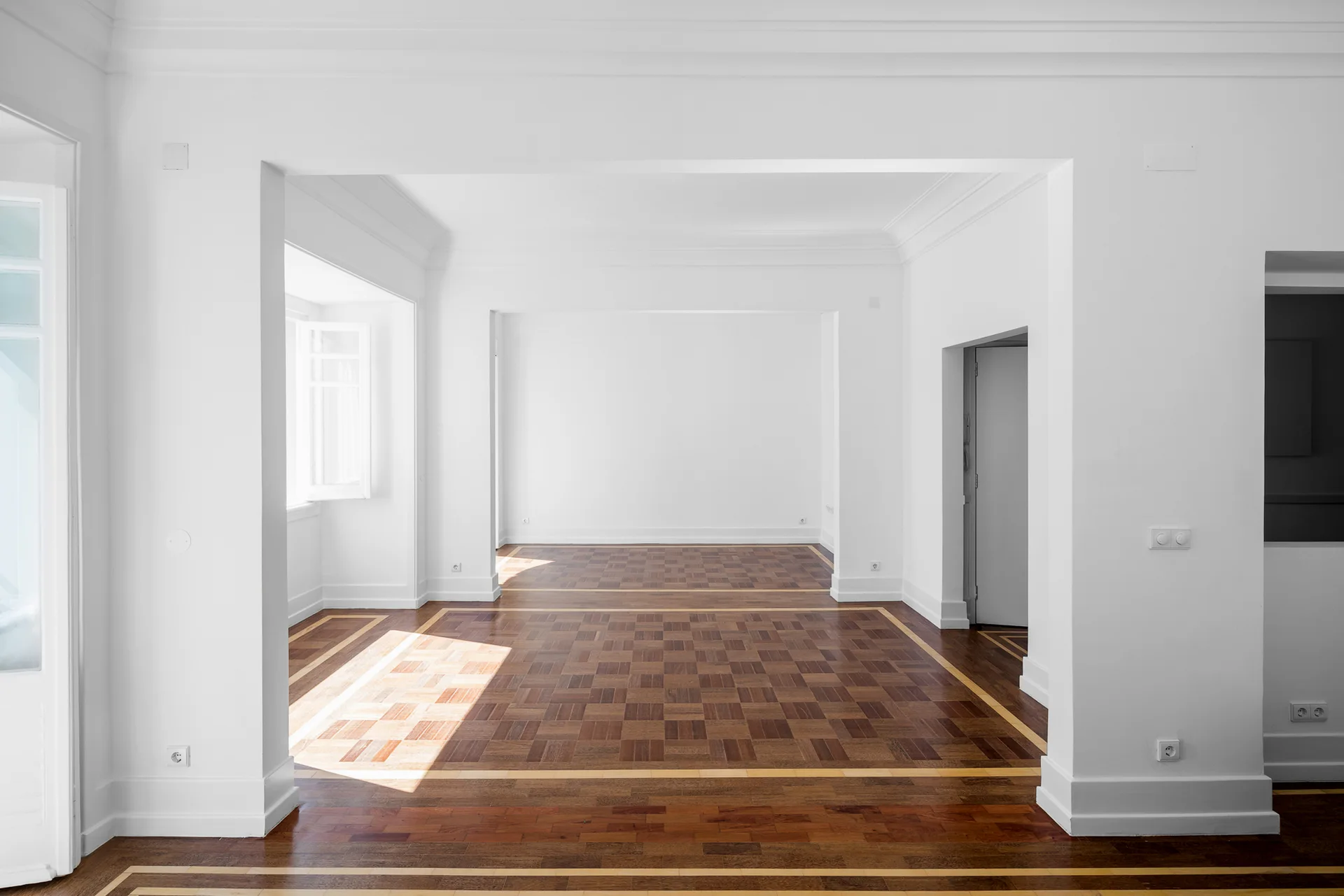 An empty white room features classical crown molding and glossy checkerboard-patterned parquet wood floors viewed through wide open doorways.