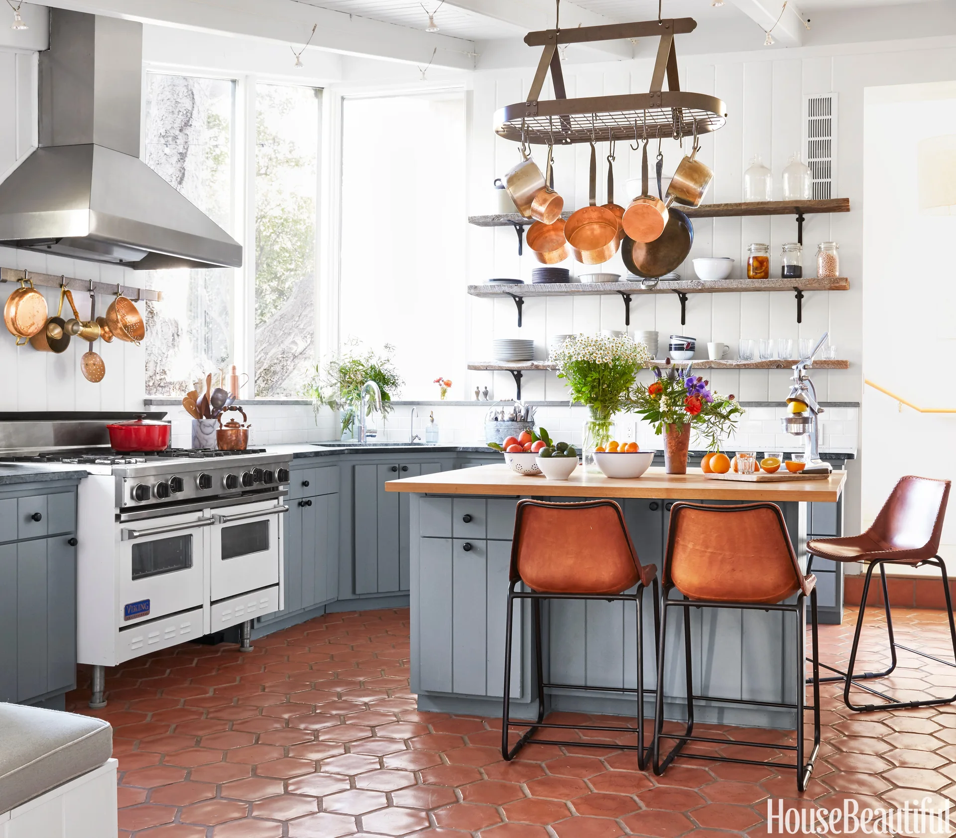 A rustic kitchen with gray cabinets, a wood-topped island with leather stools, hanging copper pots, and hexagonal terracotta floor tiles.