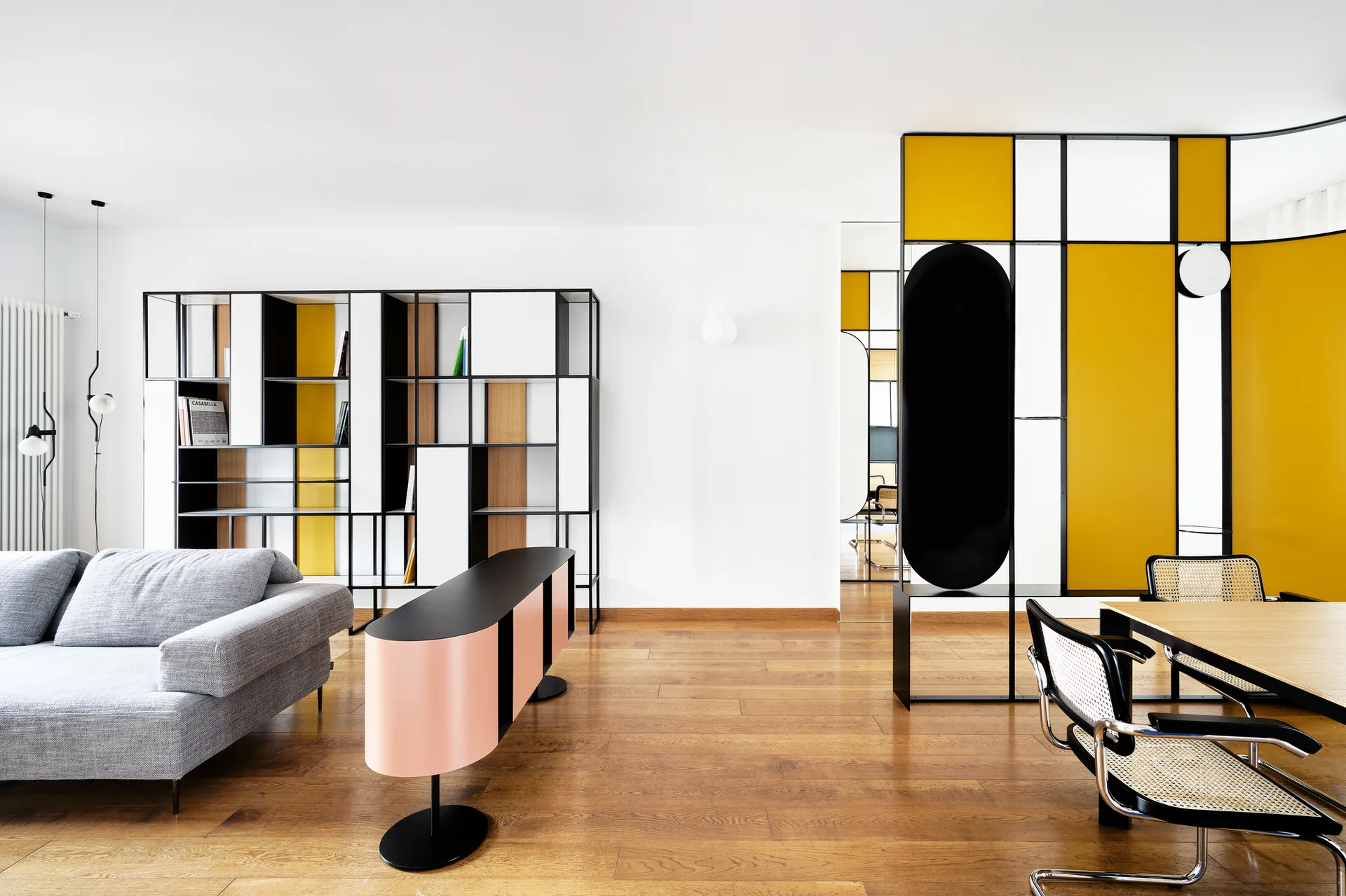 A modern living area features a grey sofa, a pink oval console, a custom wall shelving unit, and a black and yellow geometric room divider.