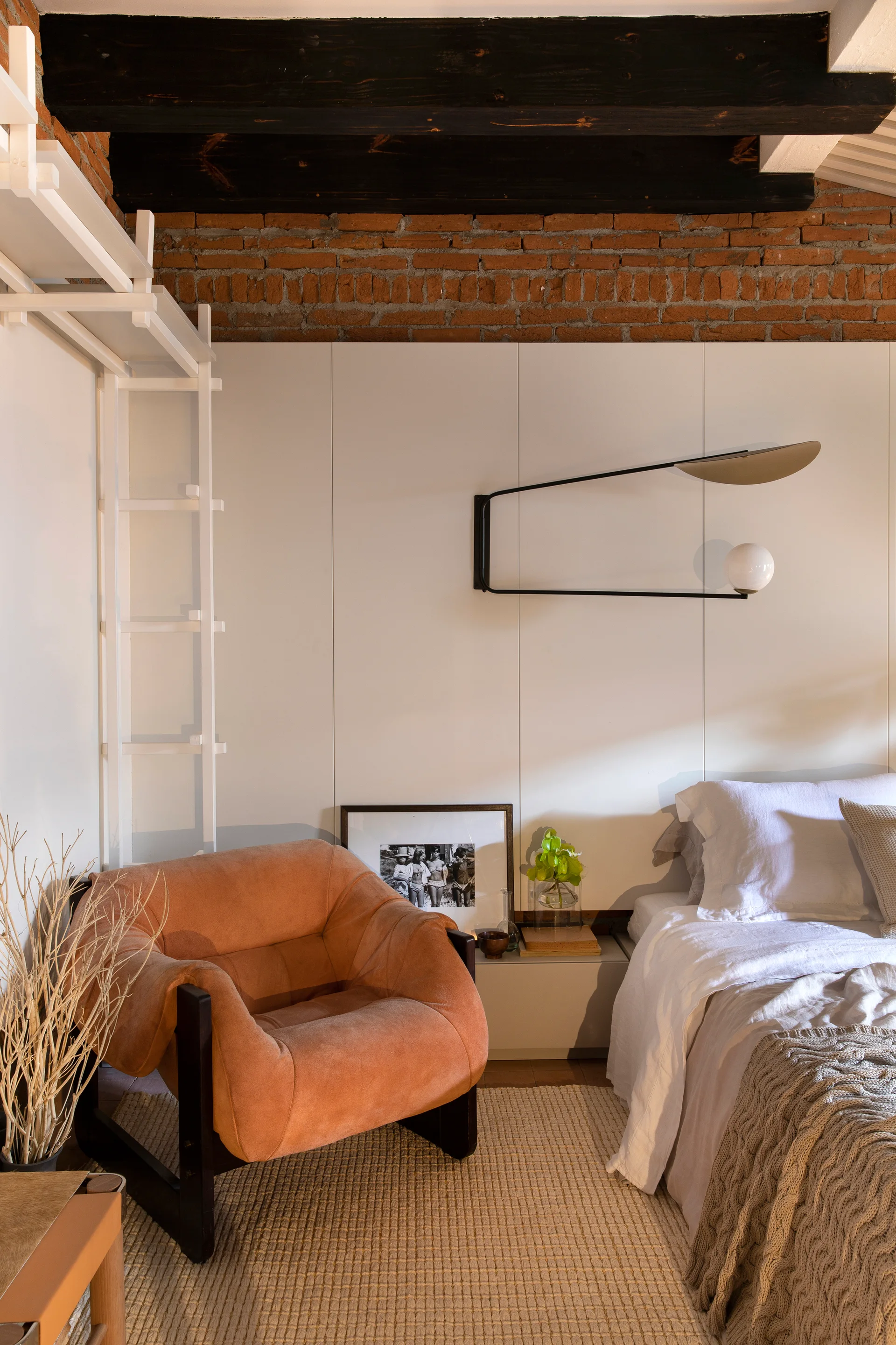 A rustic bedroom features an orange armchair next to a white bed, beneath exposed brick and dark wood ceiling beams.