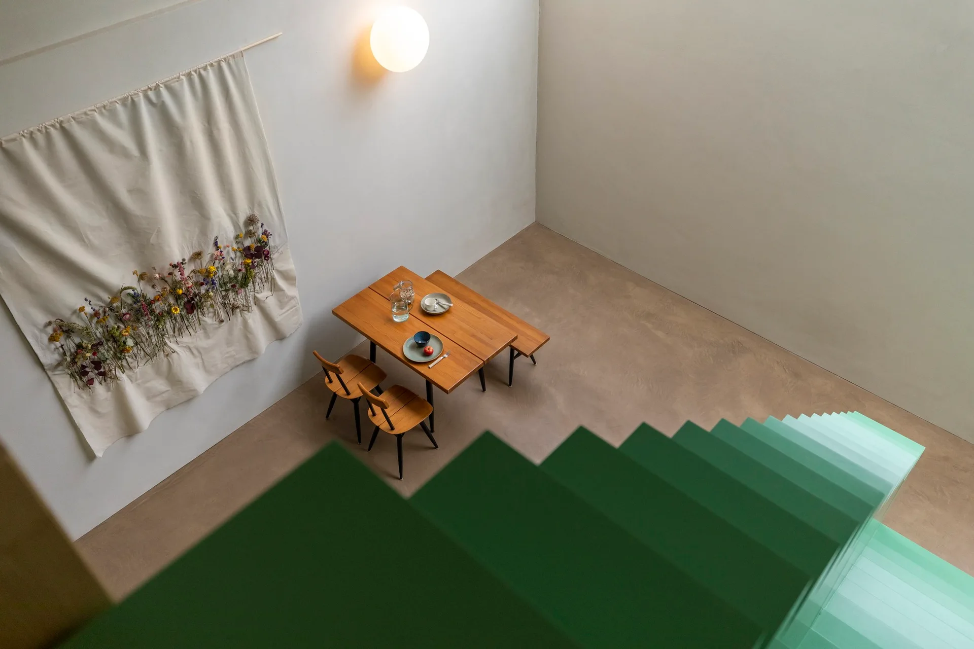 A high-angle view of a minimalist dining area featuring a wooden table, chairs, a wildflower wall tapestry, and a green gradient staircase.
