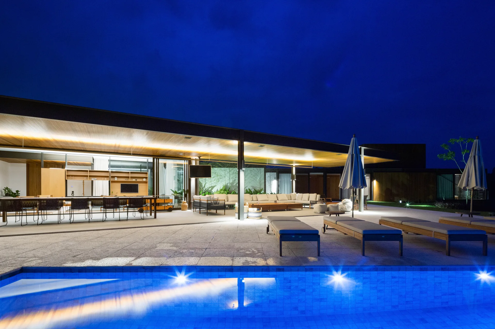 A modern outdoor patio at night features an illuminated blue swimming pool, sun loungers, and a covered lounge area with a wood-slatted ceiling.