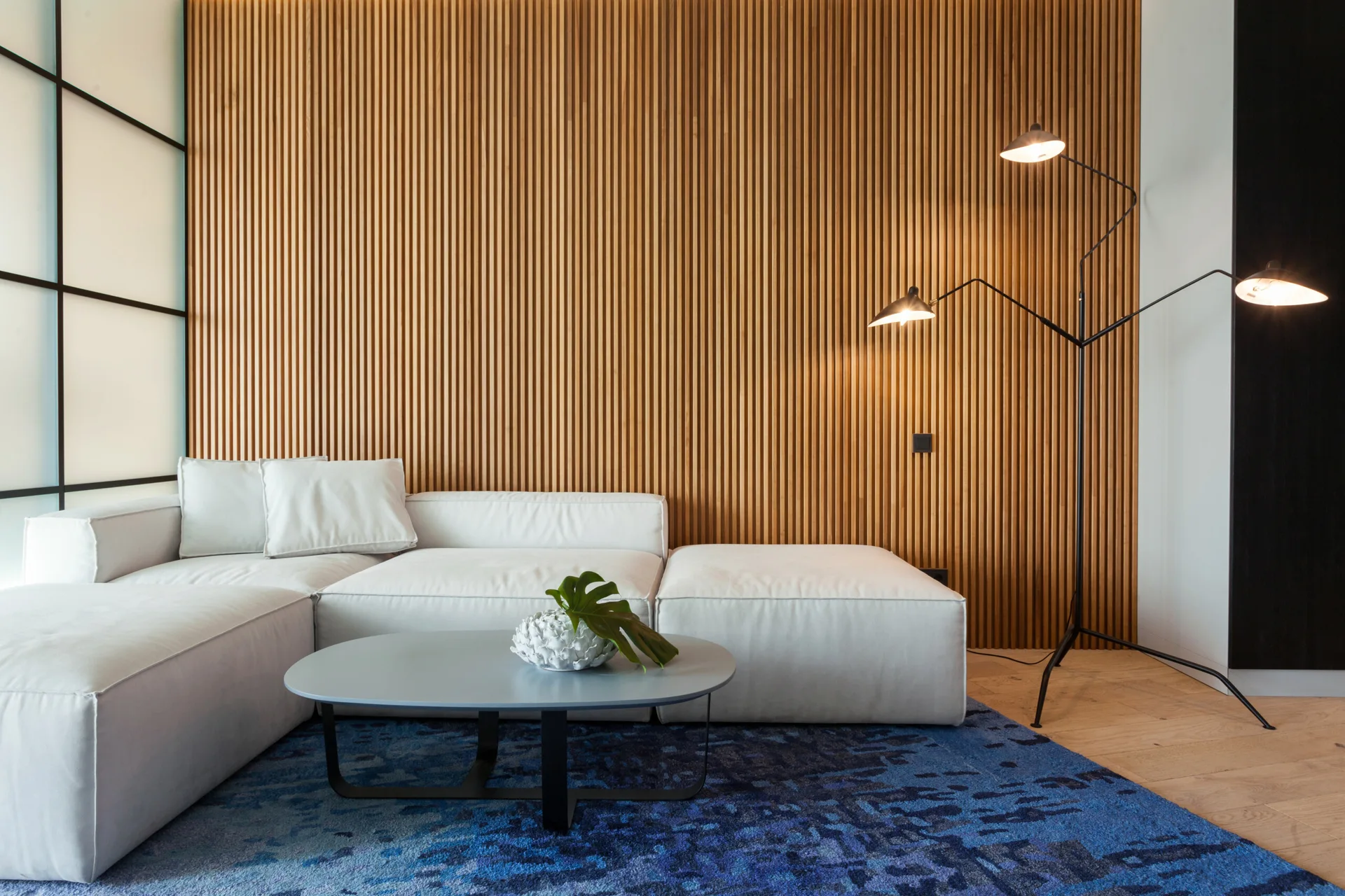 A modern living room features a white modular sectional on a blue patterned rug, set against a vertical wood slat accent wall.