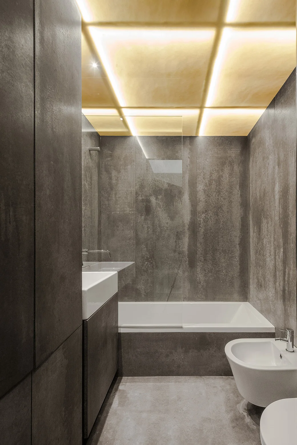 A minimalist bathroom features grey stone wall panels, a white bathtub with a glass screen, and an illuminated warm gold grid ceiling.
