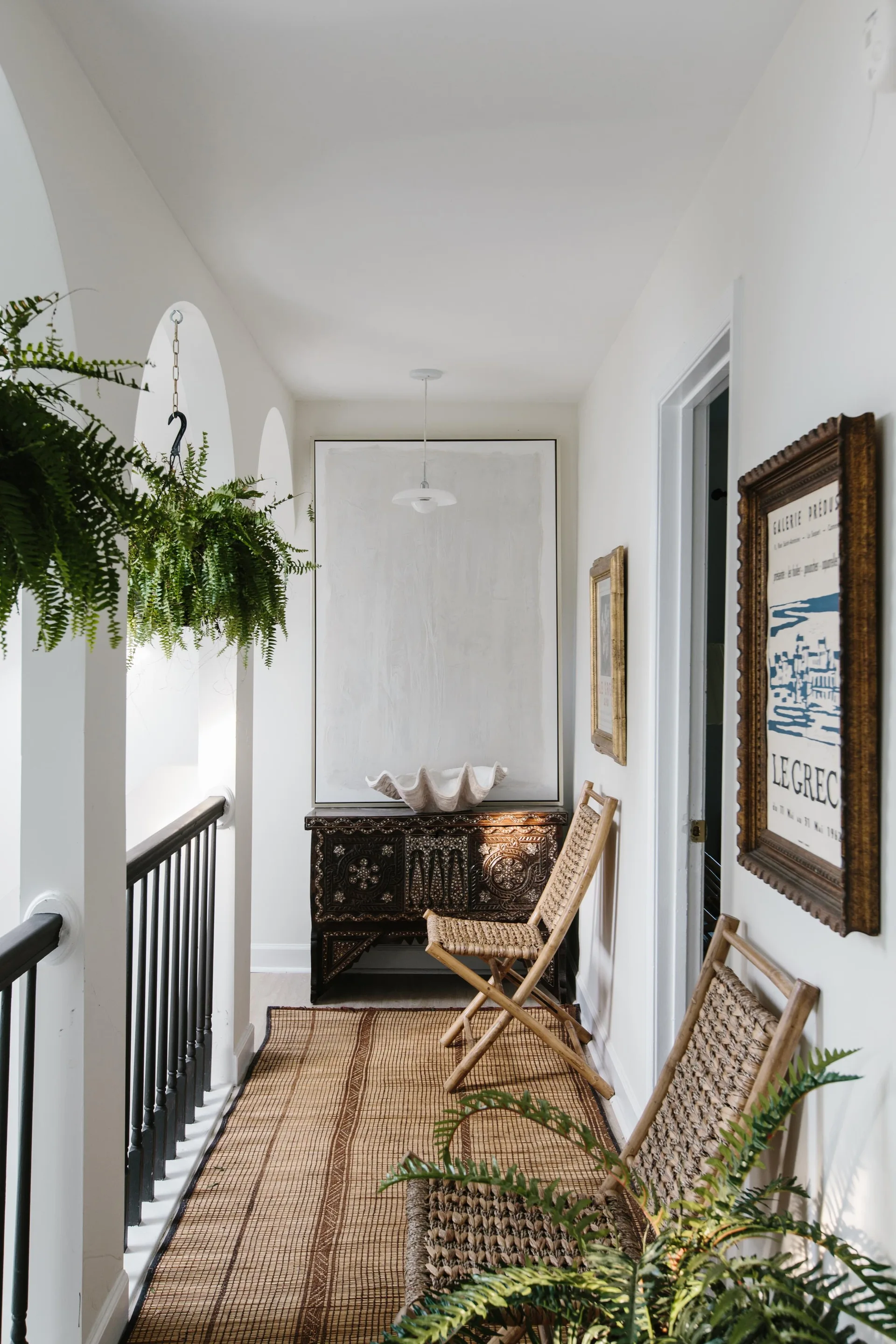 A white hallway landing features a dark carved wood console table, two woven folding chairs, a natural fiber runner, and hanging plants.