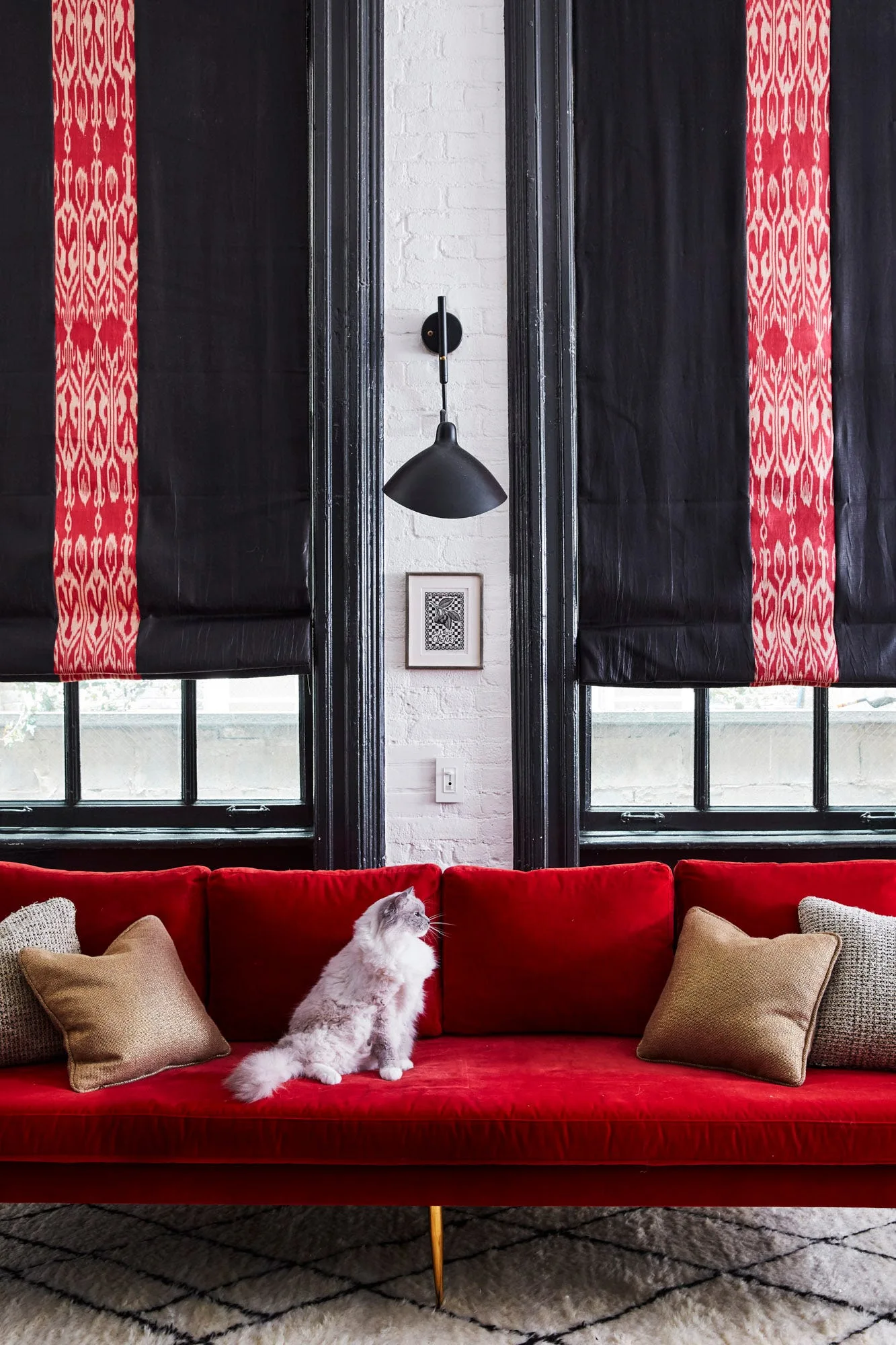 A long red velvet sofa sits before two large black-framed windows with patterned black and red roman shades on a white painted brick wall.