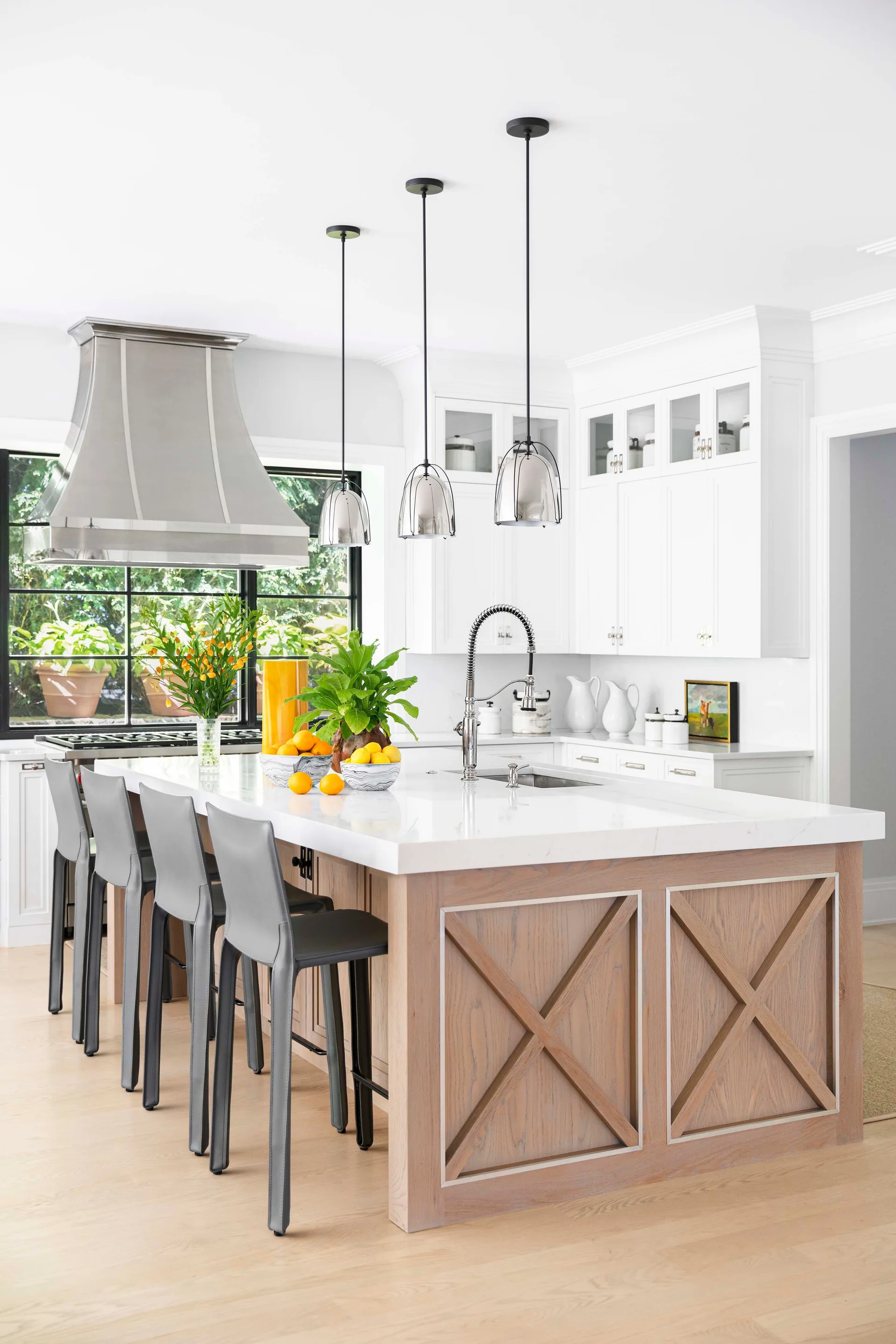 A bright kitchen features a white-topped wood island with X-brace detailing, grey bar stools, white cabinetry, and three pendant lights.