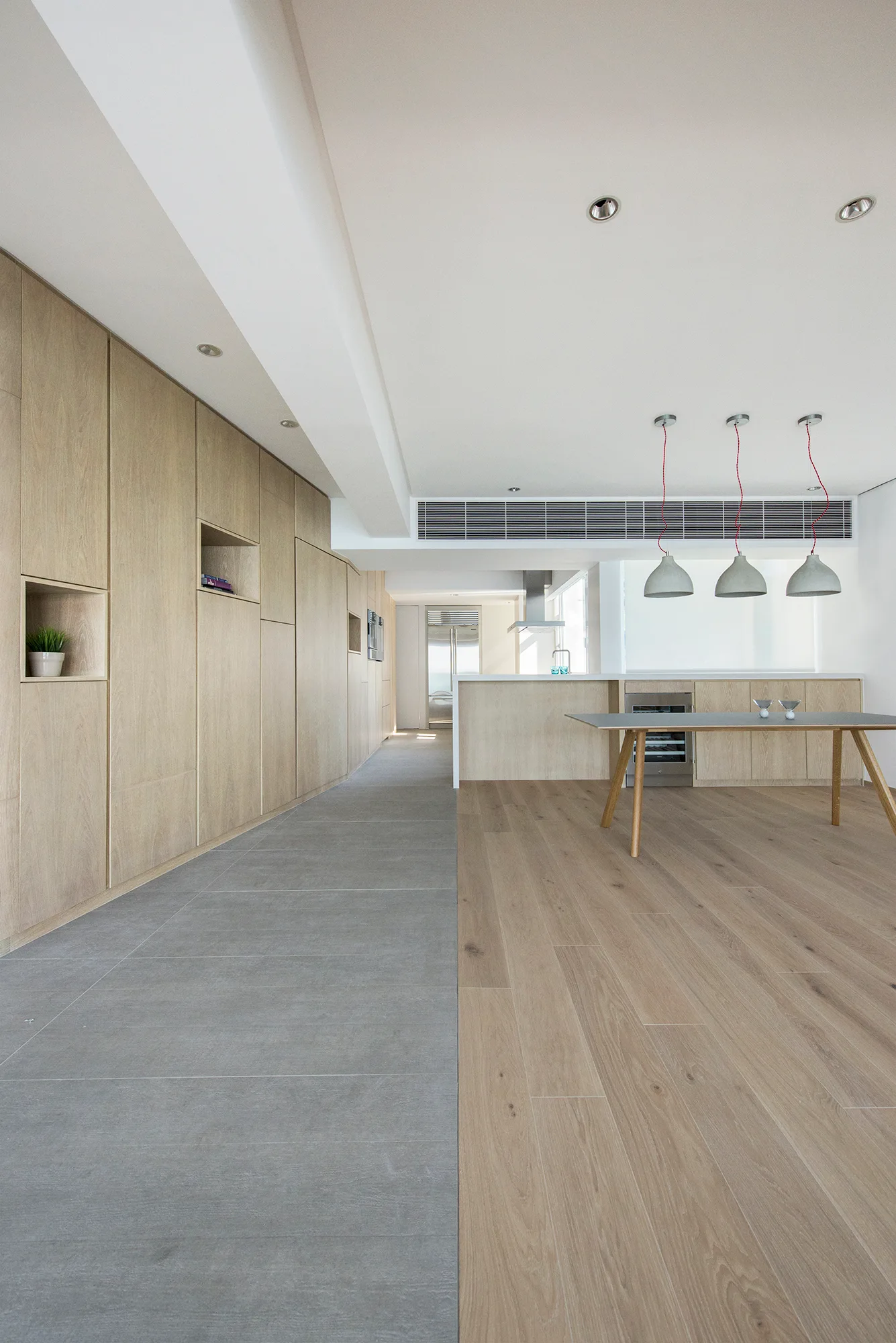 A modern kitchen space features a wall of light oak cabinetry, a grey-topped dining table, and a floor divided between grey tiles and wood planks.