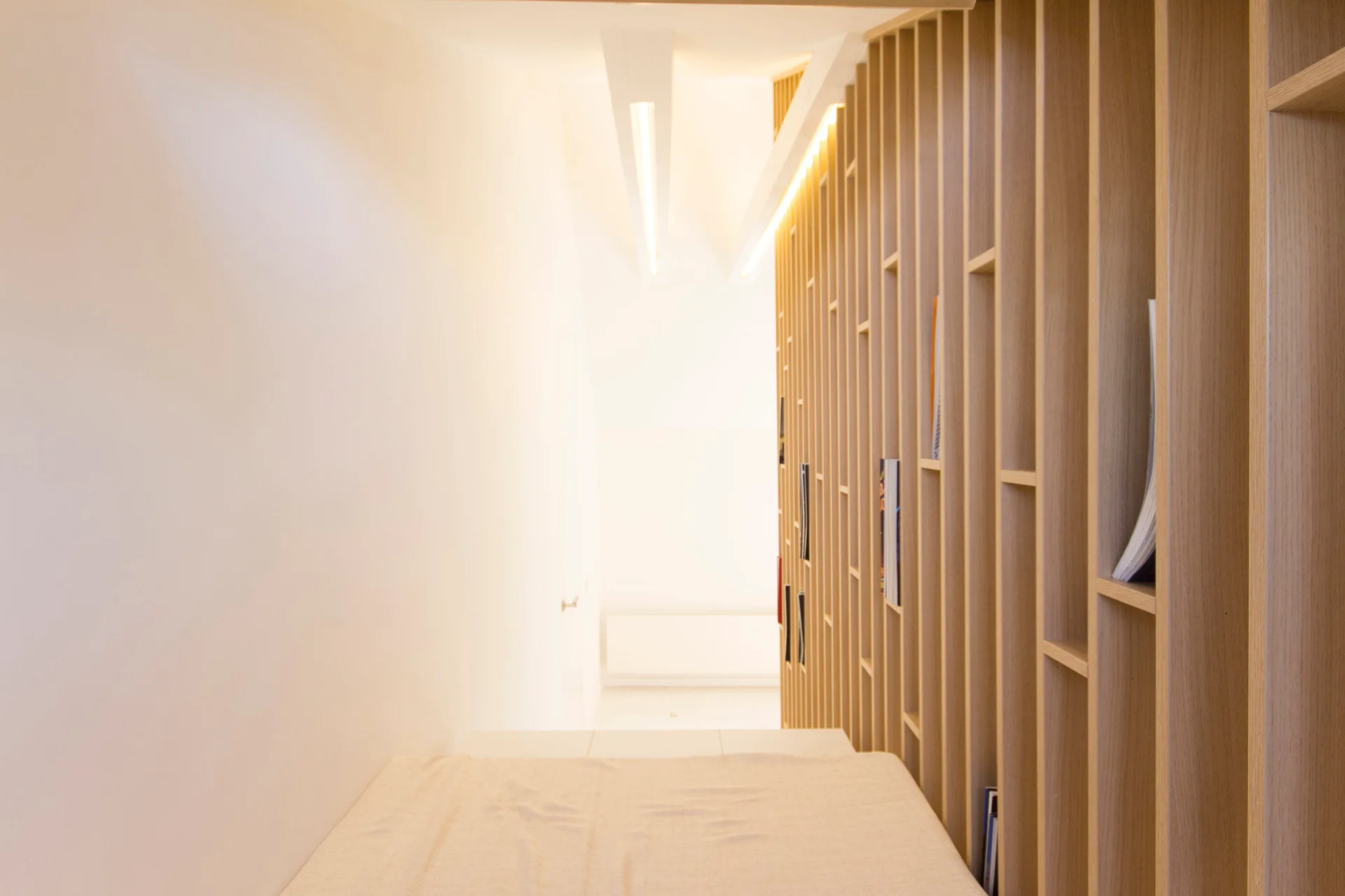 A modern hallway featuring a white painted wall opposite a light wood vertical slat wall with integrated bookshelves and recessed ceiling lights.