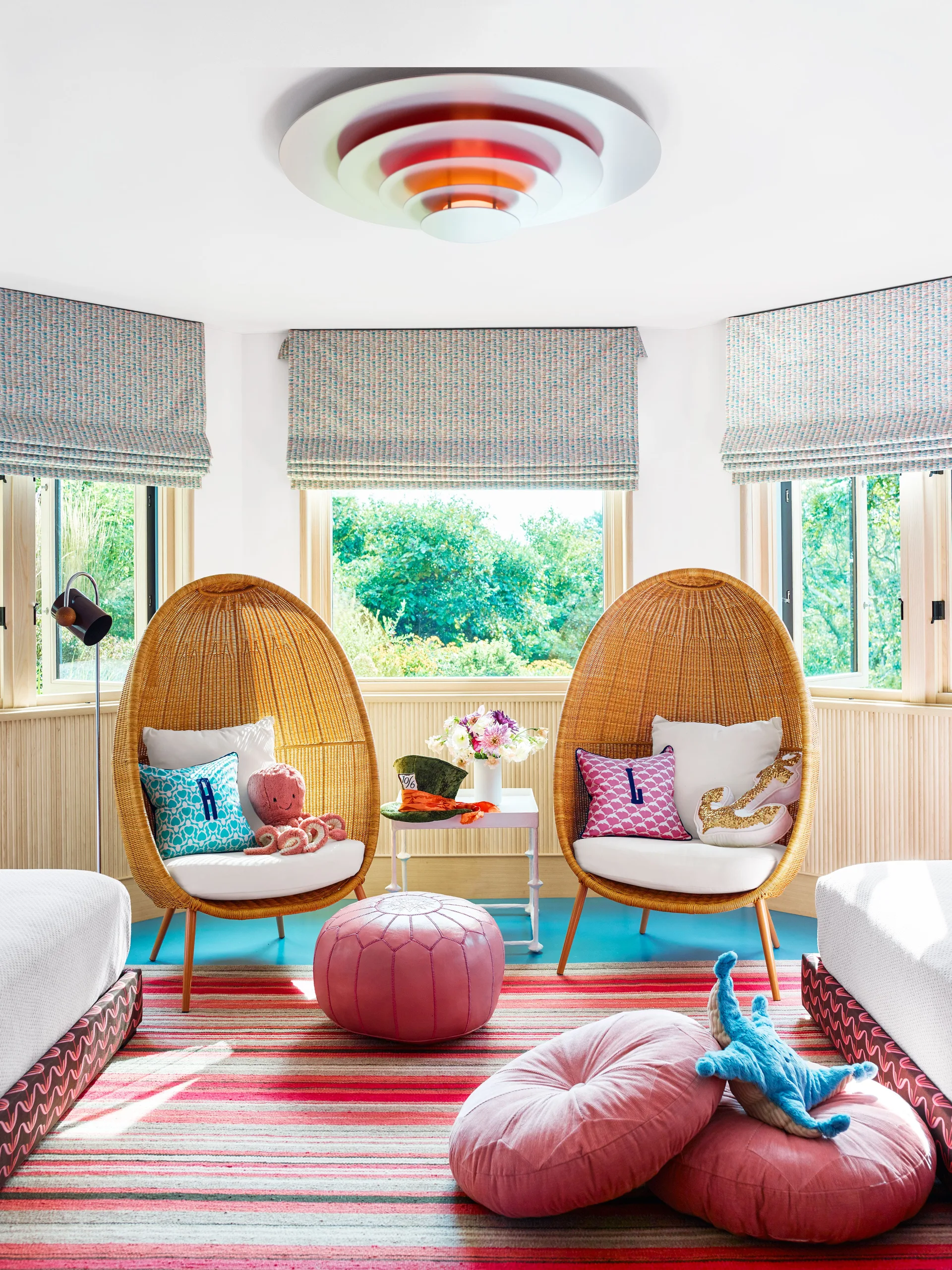 Two woven rattan egg chairs sit in a bright bay window alcove with patterned roman shades, a colorful striped rug, and pink floor cushions.