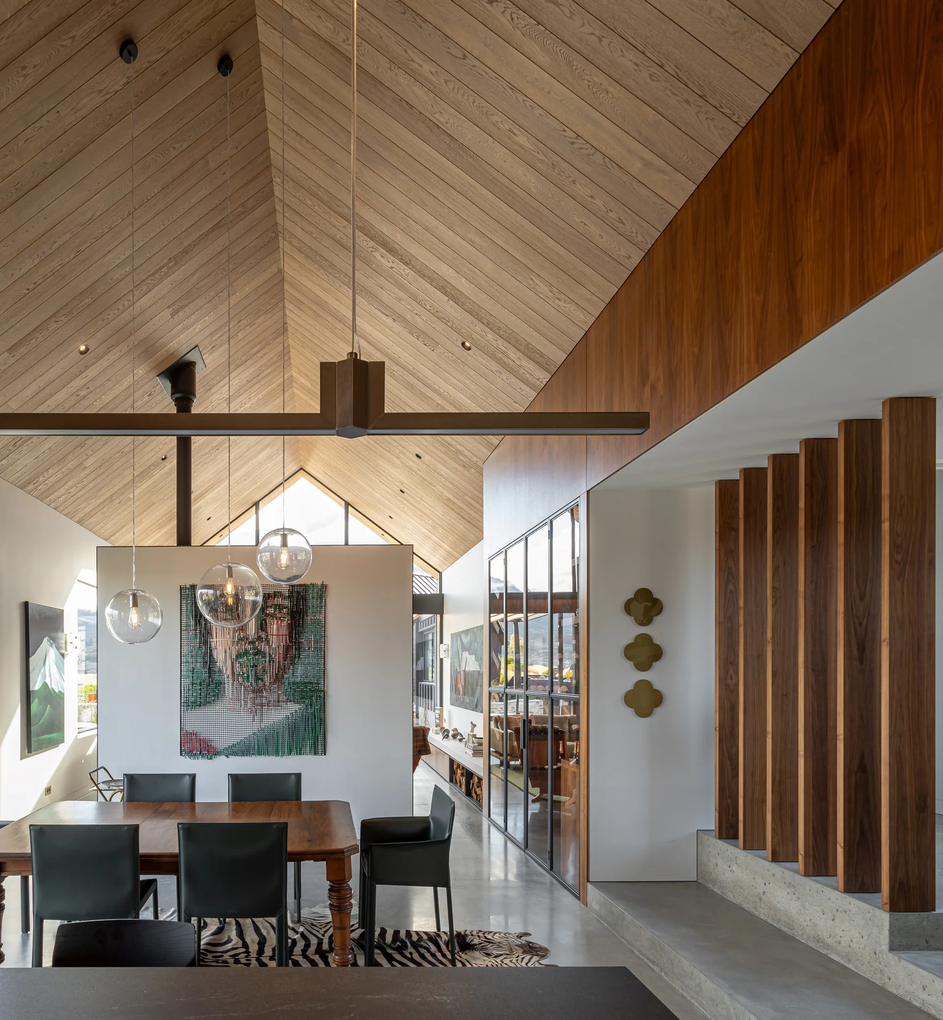 A modern dining room features a high vaulted wood ceiling, a wooden table with black chairs on an animal print rug, and a glass-paneled divider.