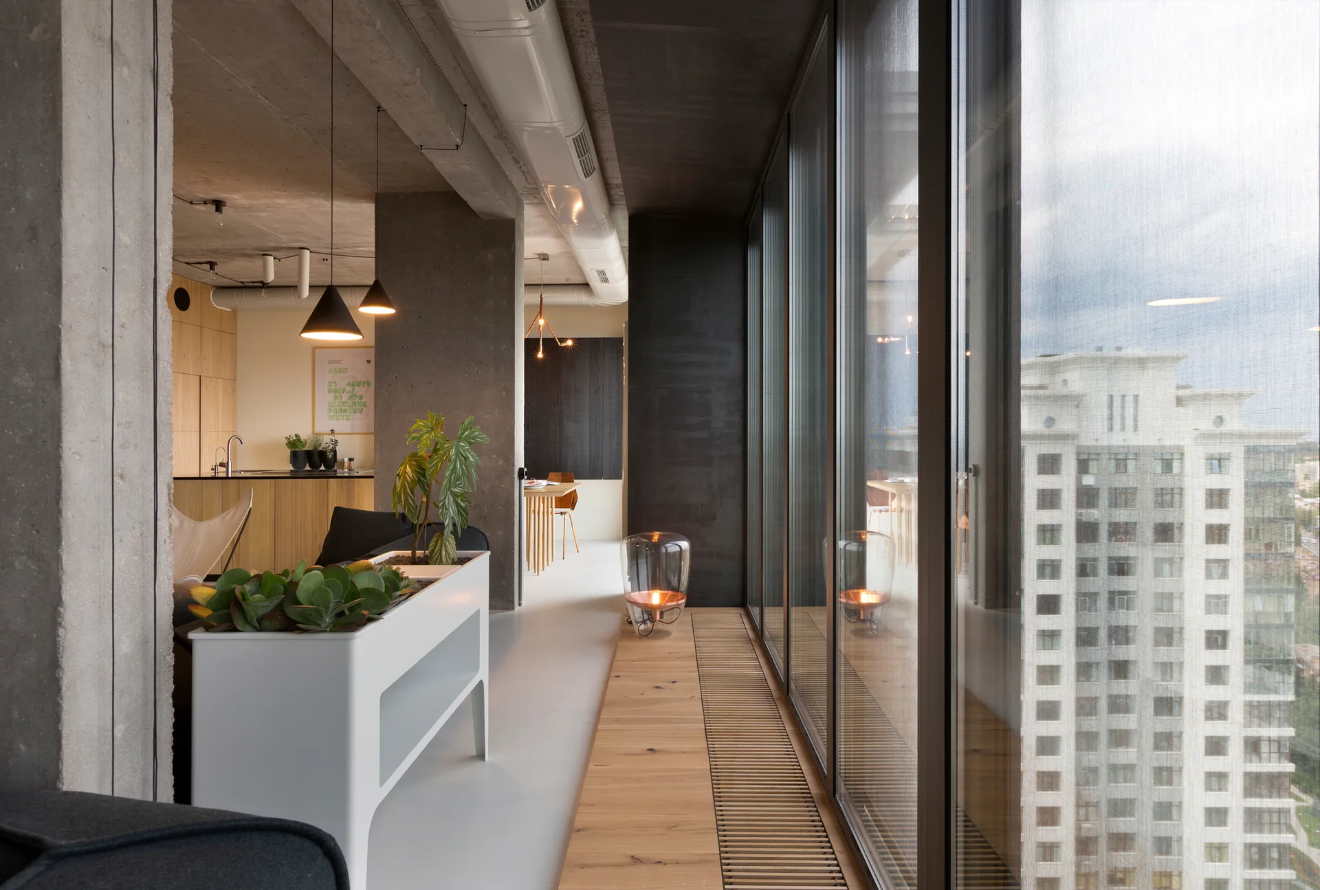 An industrial apartment interior features raw concrete ceilings, large floor-to-ceiling windows, a white planter, and a wood slatted floor grate.
