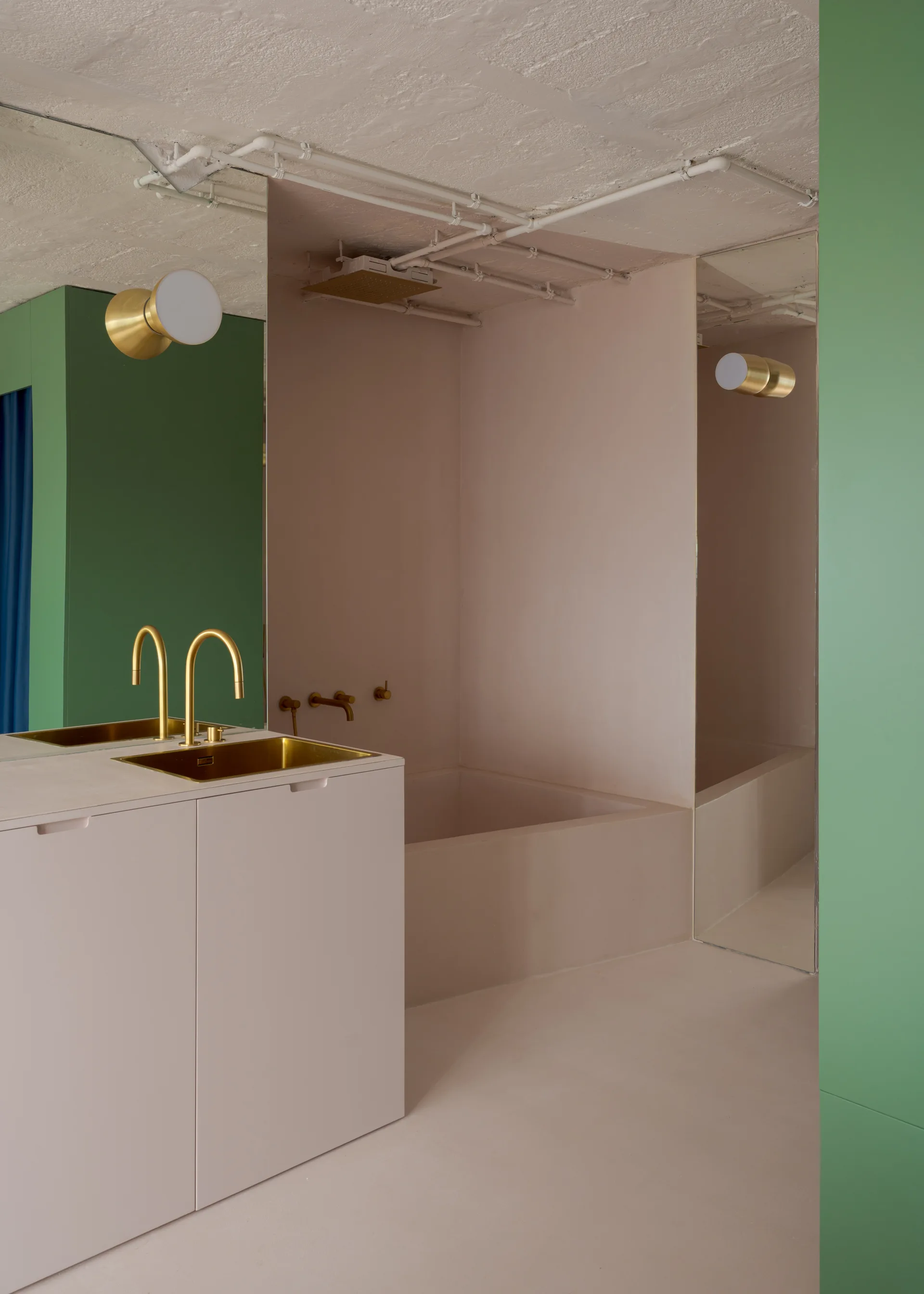 A modern bathroom with a muted pink vanity and matching bathtub alcove, contrasted by bold green wall panels and brass fixtures.