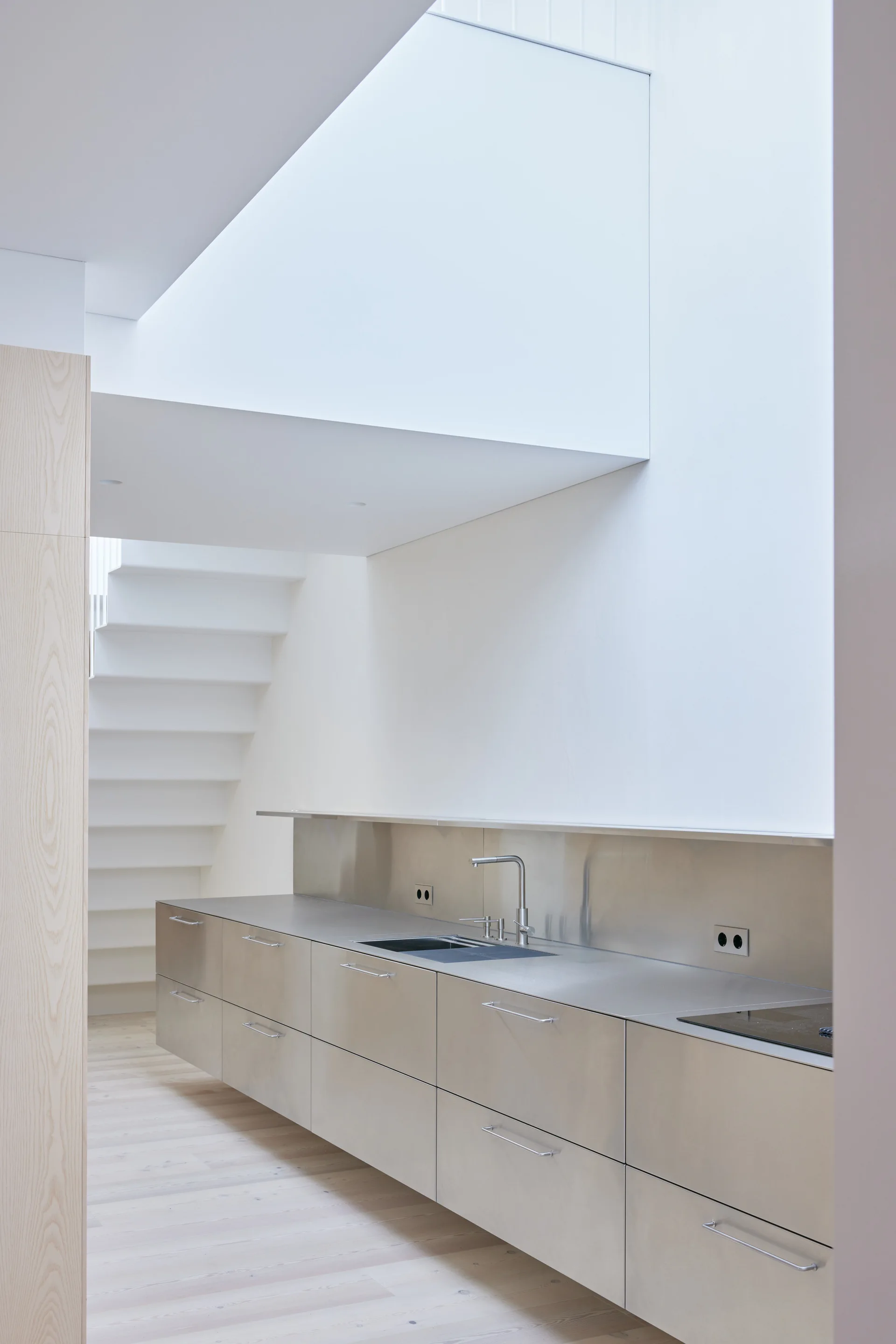 A minimalist kitchen features sleek stainless steel cabinets, a matching backsplash, a light wood floor, and a white staircase in the background.